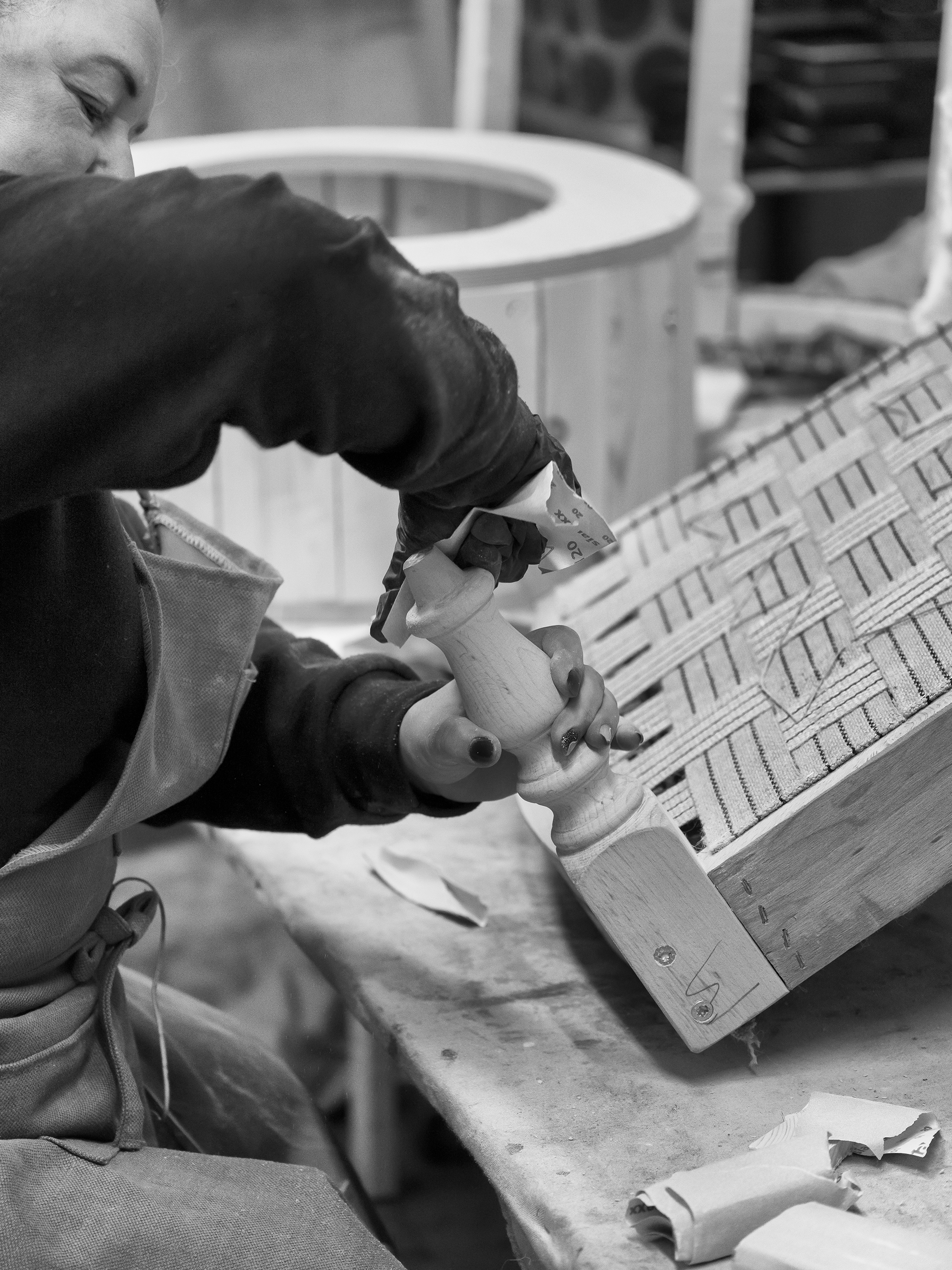 Person carving wood with a chisel, wearing an apron. The workspace has a wooden object with a lattice pattern. Black and white image.