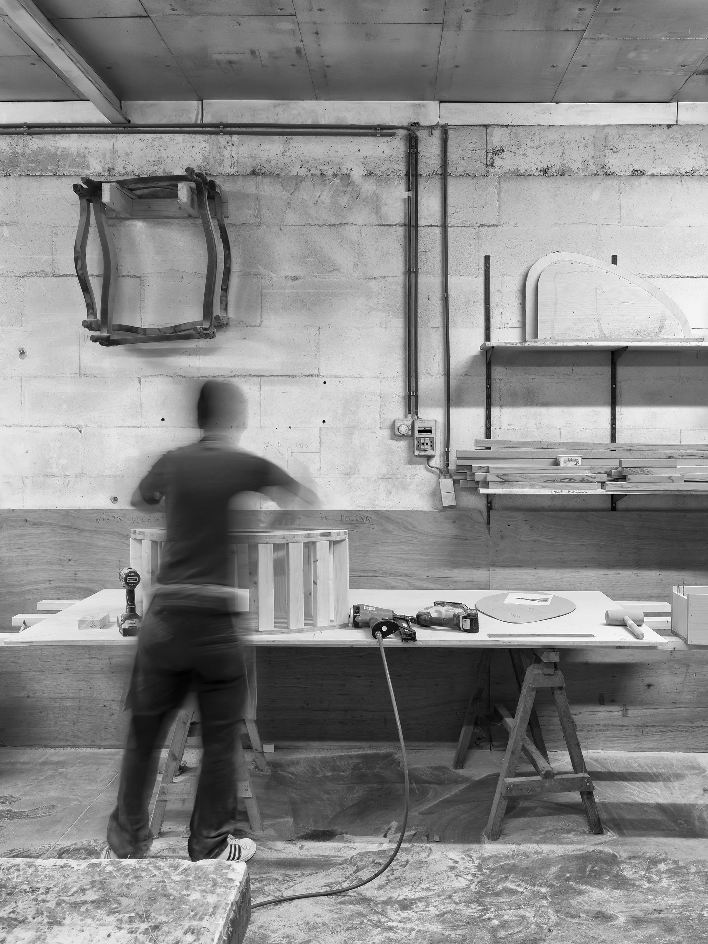 Person working on a wooden barrel in a workshop, with tools on the table and shelves in the background. Black and white image.