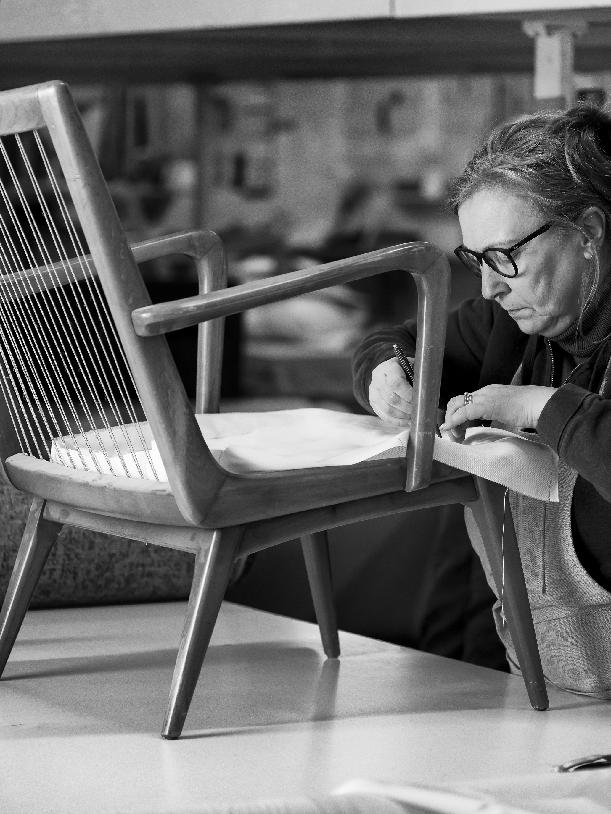 A woman wearing glasses works on upholstering a wooden chair, using tools at a table in a workshop.