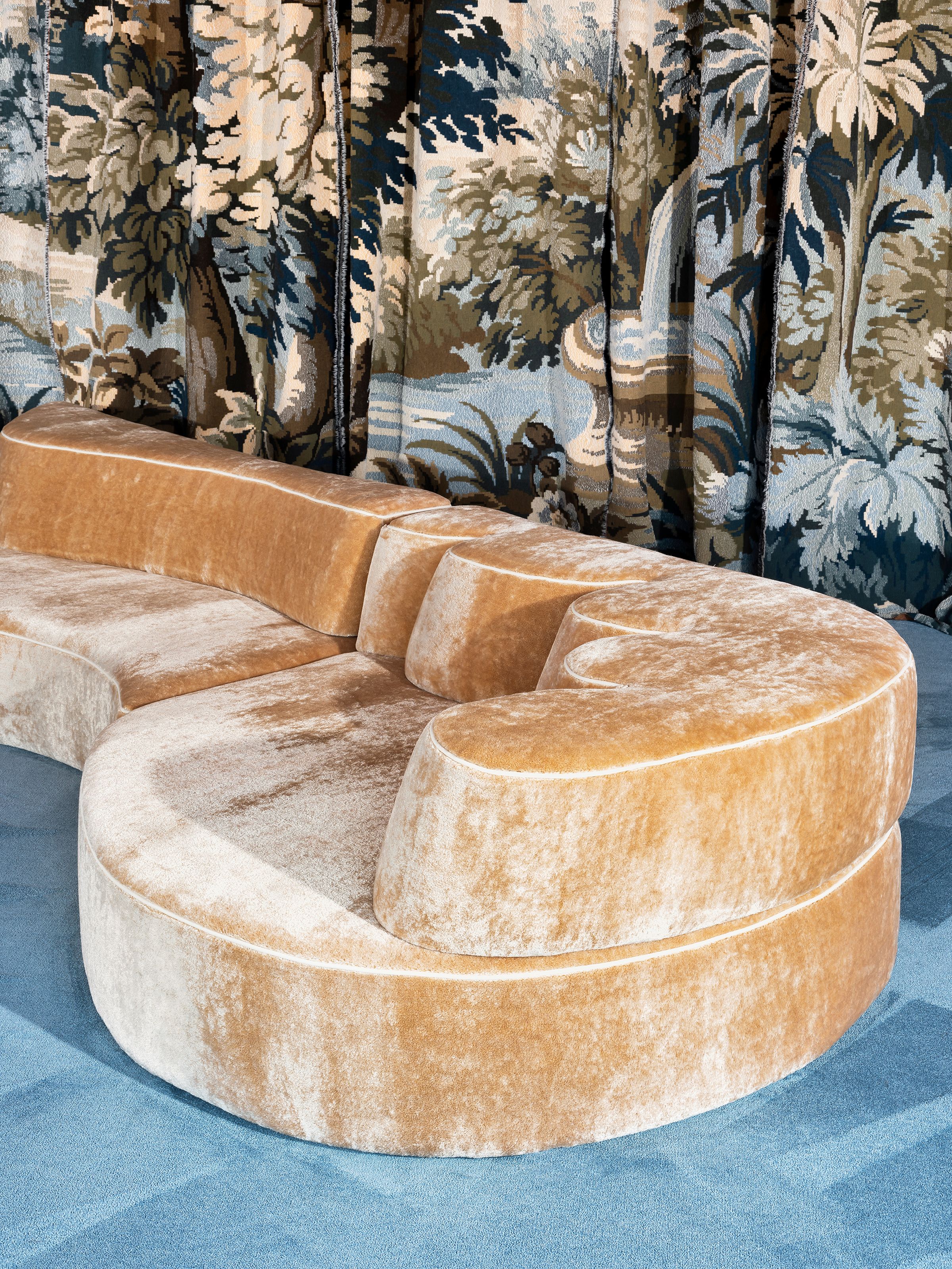 Detail of the Comporta modular sofa in plush golden velvet, showcasing its sculptural curved form and tiered backrest, placed on a blue carpet with a forest-themed tapestry curtain in the background.