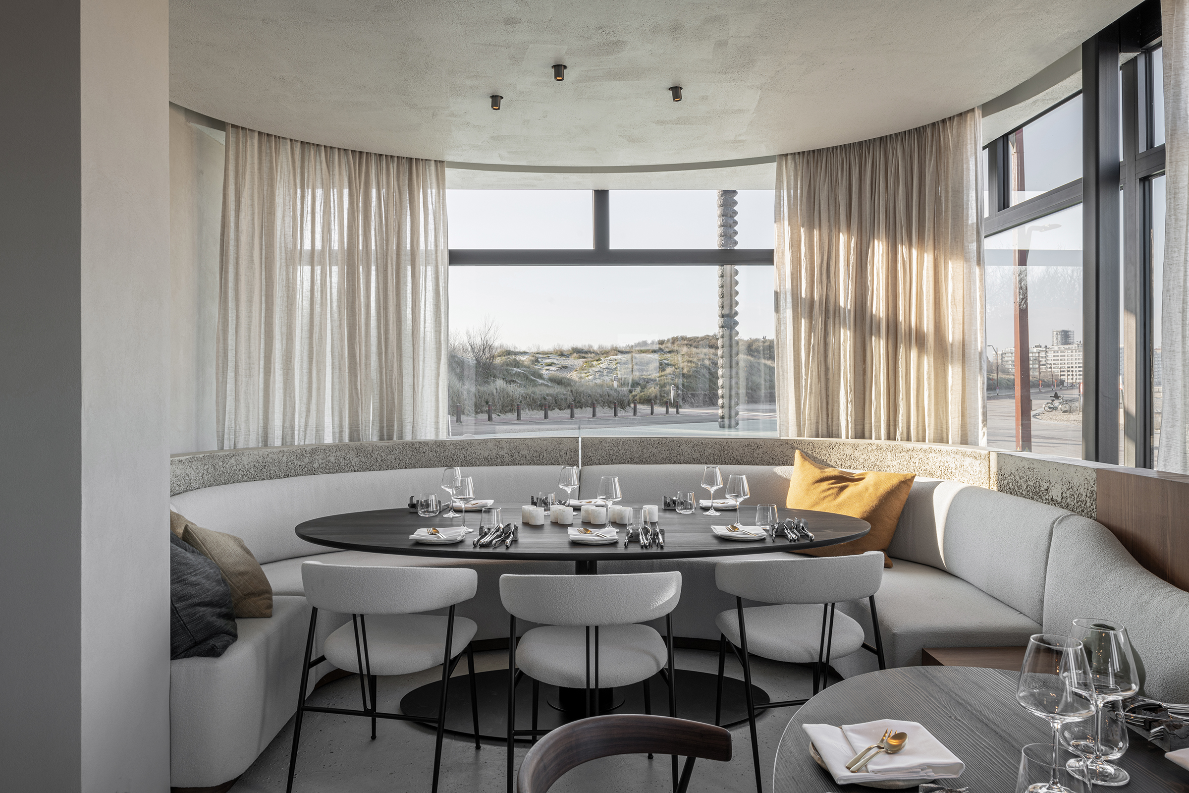 Curved banquette seating in light fabric surrounds a dark oval dining table set with glassware and cutlery, placed in a modern restaurant with floor-to-ceiling windows overlooking sand dunes and city views.