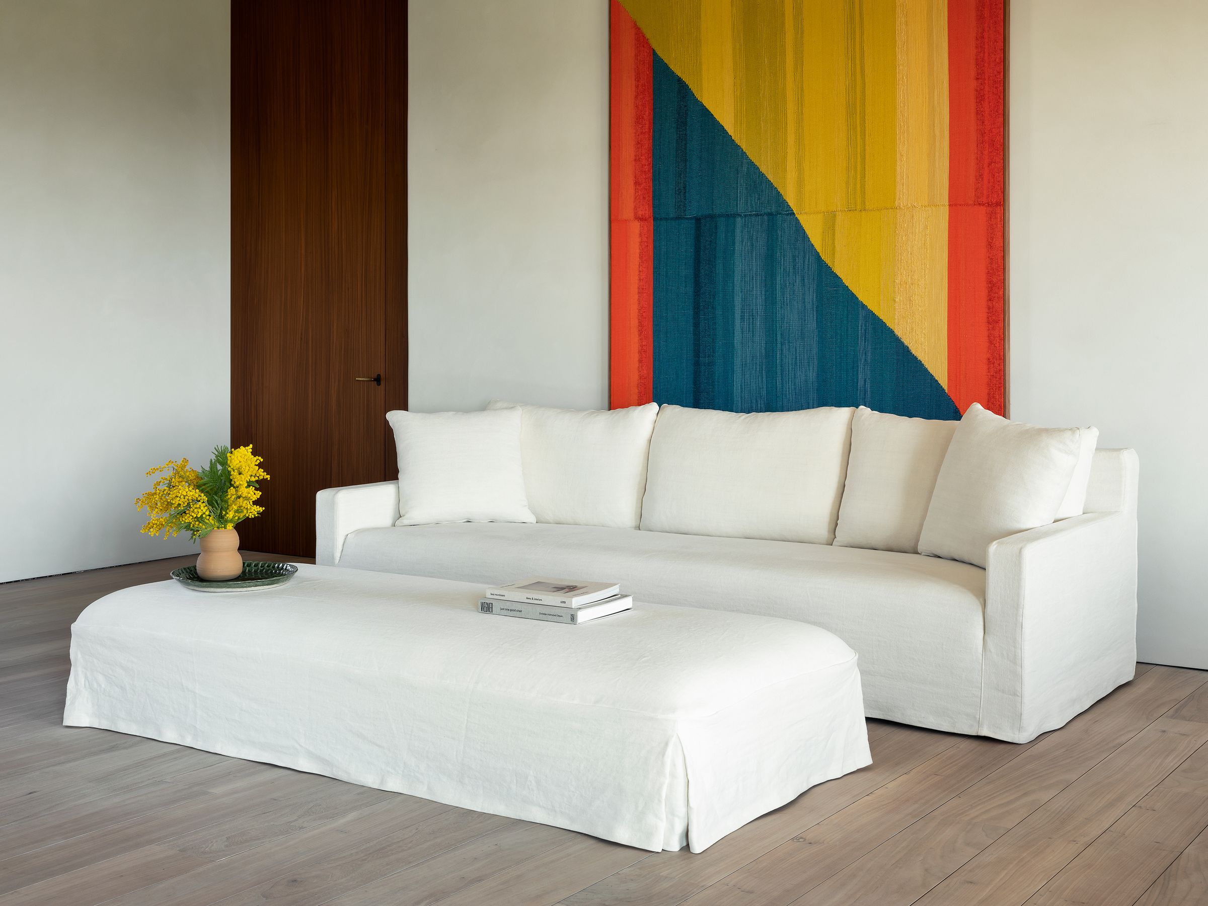 Charlie sofa in off-white fabric with clean lines and five matching cushions, paired with a large matching ottoman used as a coffee table, styled with books and a vase of yellow flowers, set against a minimalist interior with light wooden floors and a colorful geometric wall tapestry.