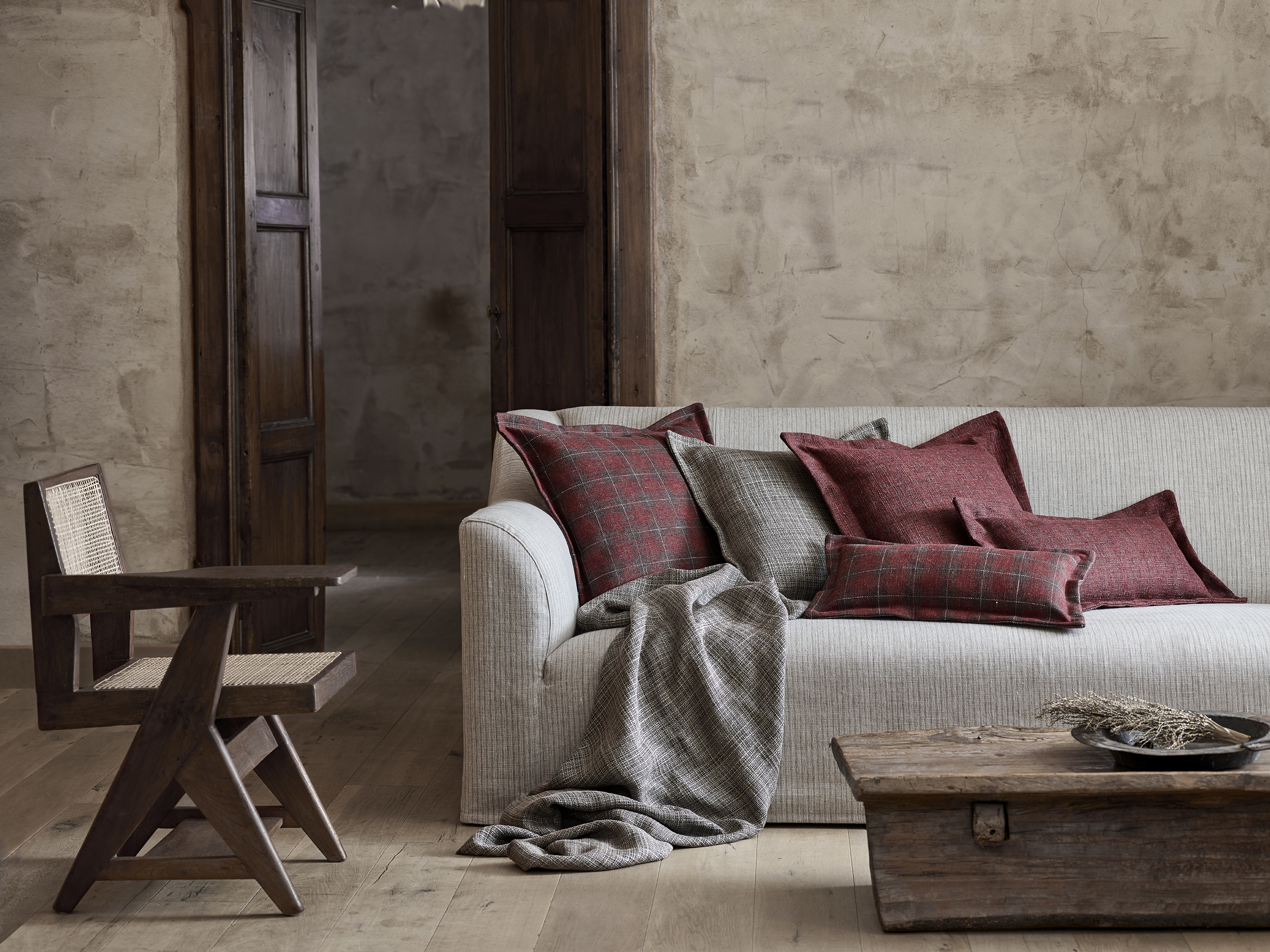 Rustic living room featuring the Toscana fixed seat sofa by Collett & Victor in a neutral fabric, styled with red and grey patterned cushions, a wooden armchair, and a vintage coffee table against textured plaster walls.