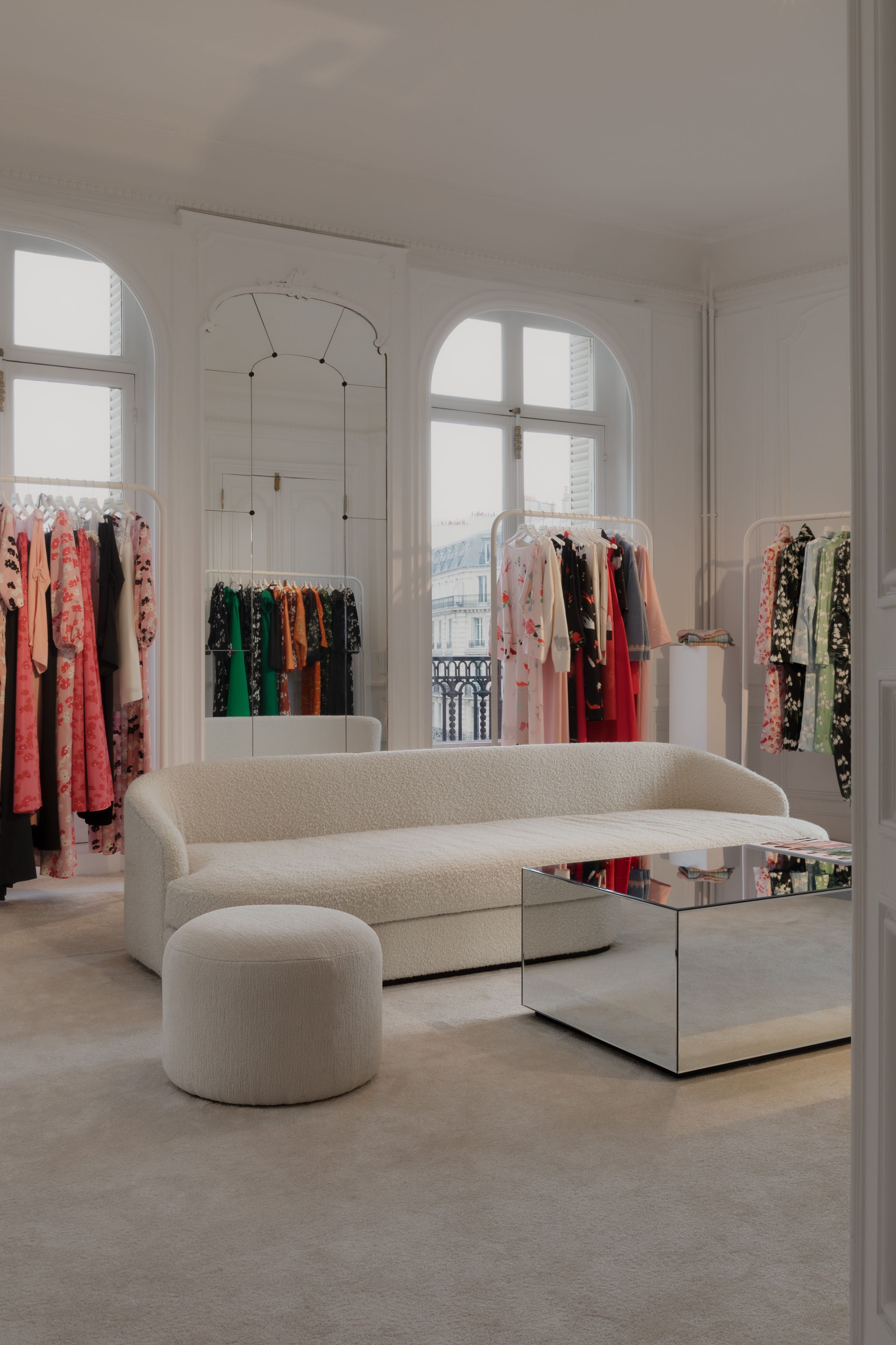 Elegant fashion showroom with a soft cream boucle sofa, round ottoman, and mirrored coffee table, surrounded by colorful designer garments on racks under tall arched windows.
