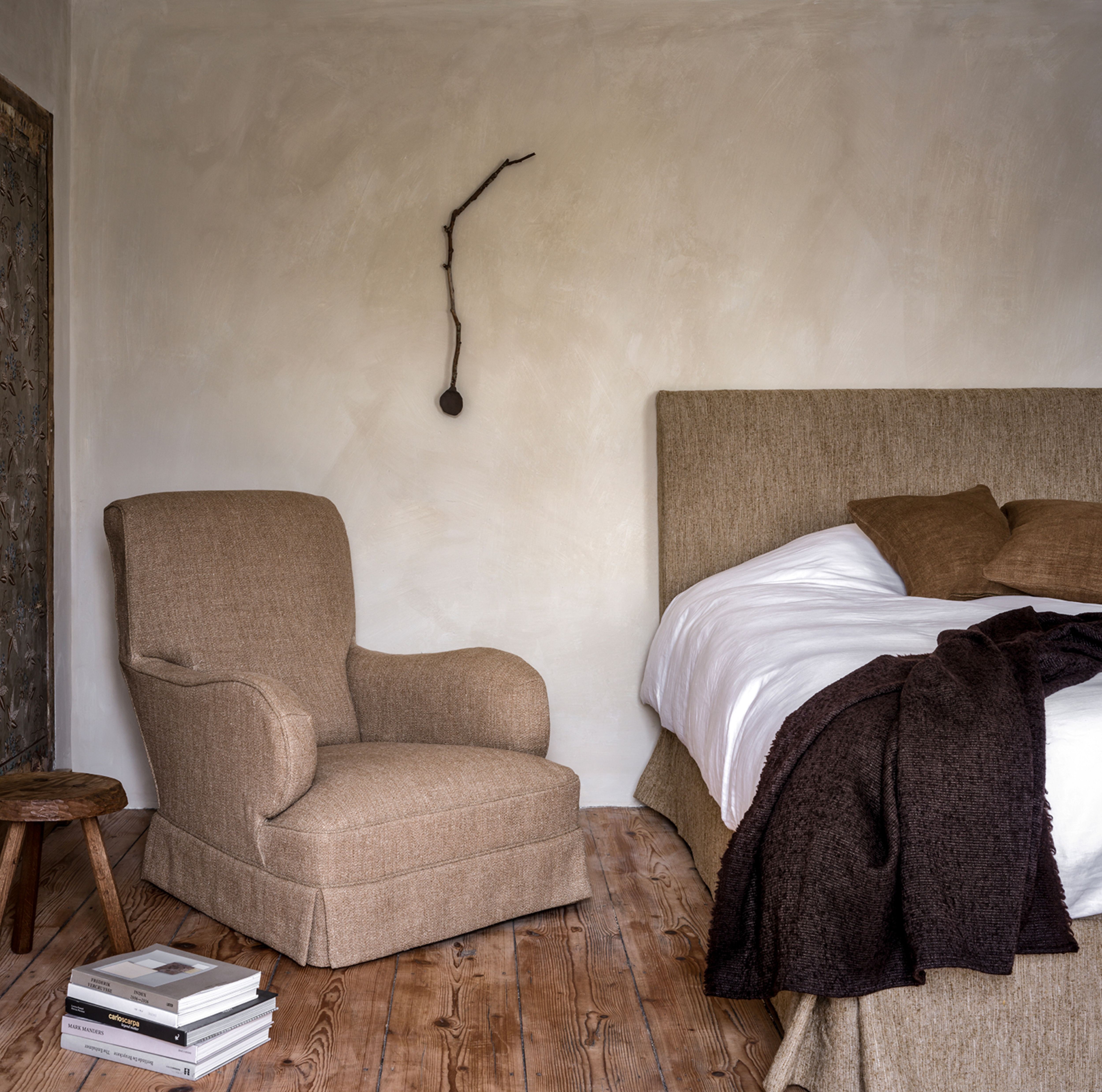 Bedroom scene with the Anaïs armchair upholstered in a warm beige fabric, placed on rustic wooden flooring beside a matching upholstered bed with white linens and brown pillows. A small wooden stool and a stack of books complete the cozy setting against a neutral plastered wall.