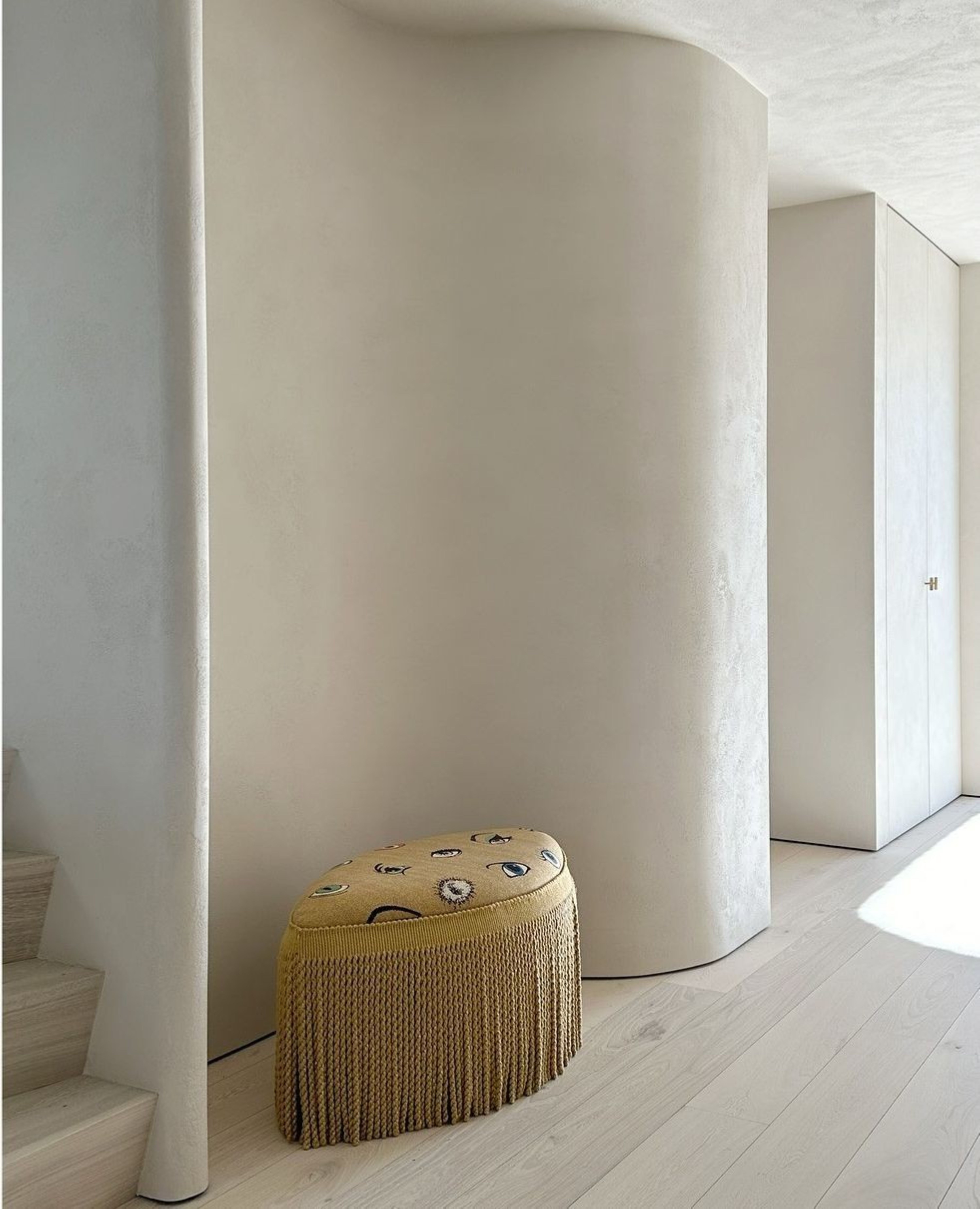Cleopatra pouf by Collett & Victor, upholstered in mustard fabric with playful embroidery and fringe, set against a minimalist curved wall.
