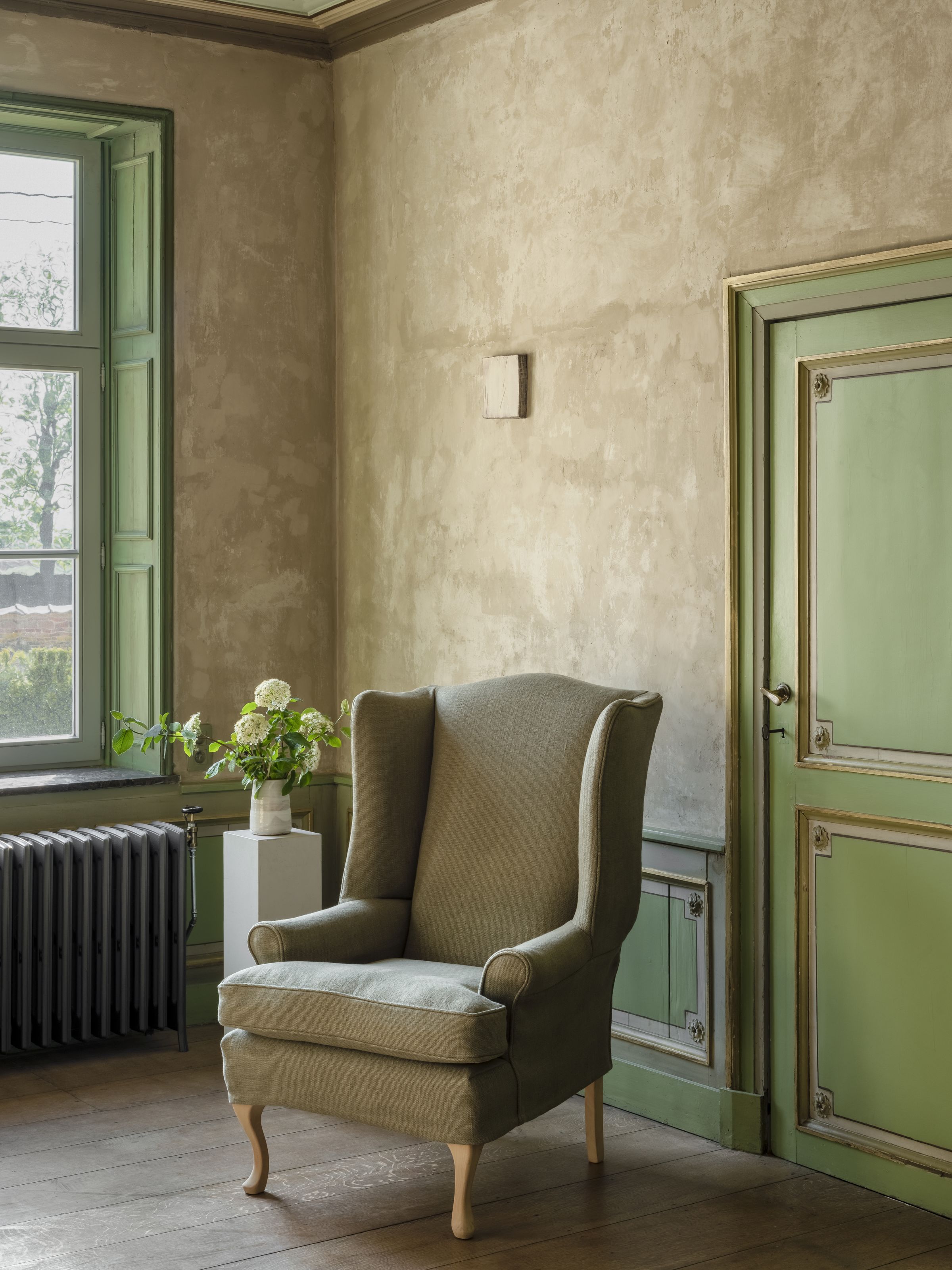 Christopher armchair in muted green upholstery with high wingback, rolled arms, and natural wooden cabriole legs. Styled in a rustic room with pale green paneled doors, a radiator, and a vase of fresh flowers by the window.