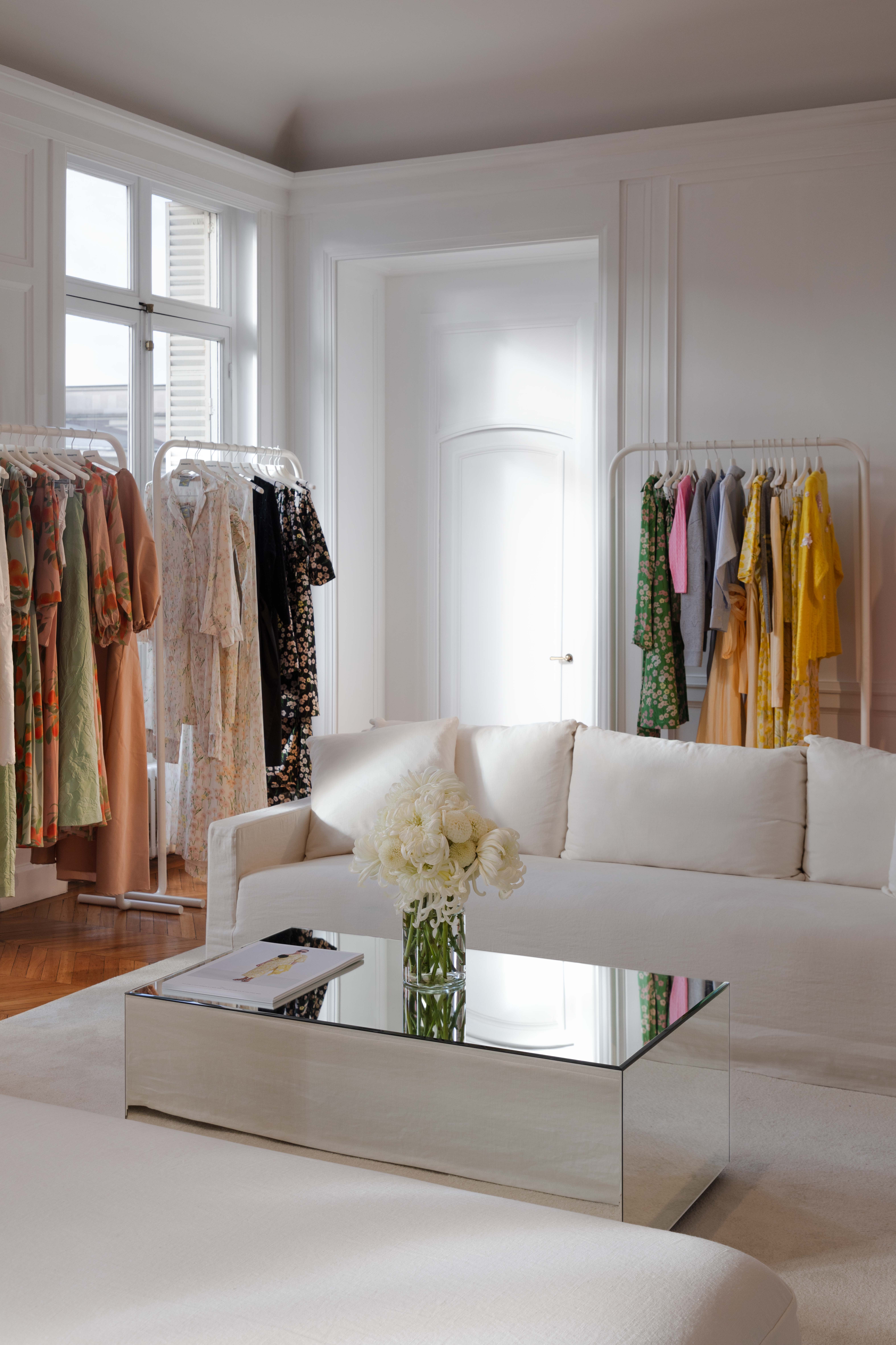 Elegant room with white sofas, a mirrored coffee table with flowers, and clothing racks displaying colorful dresses near large windows.
