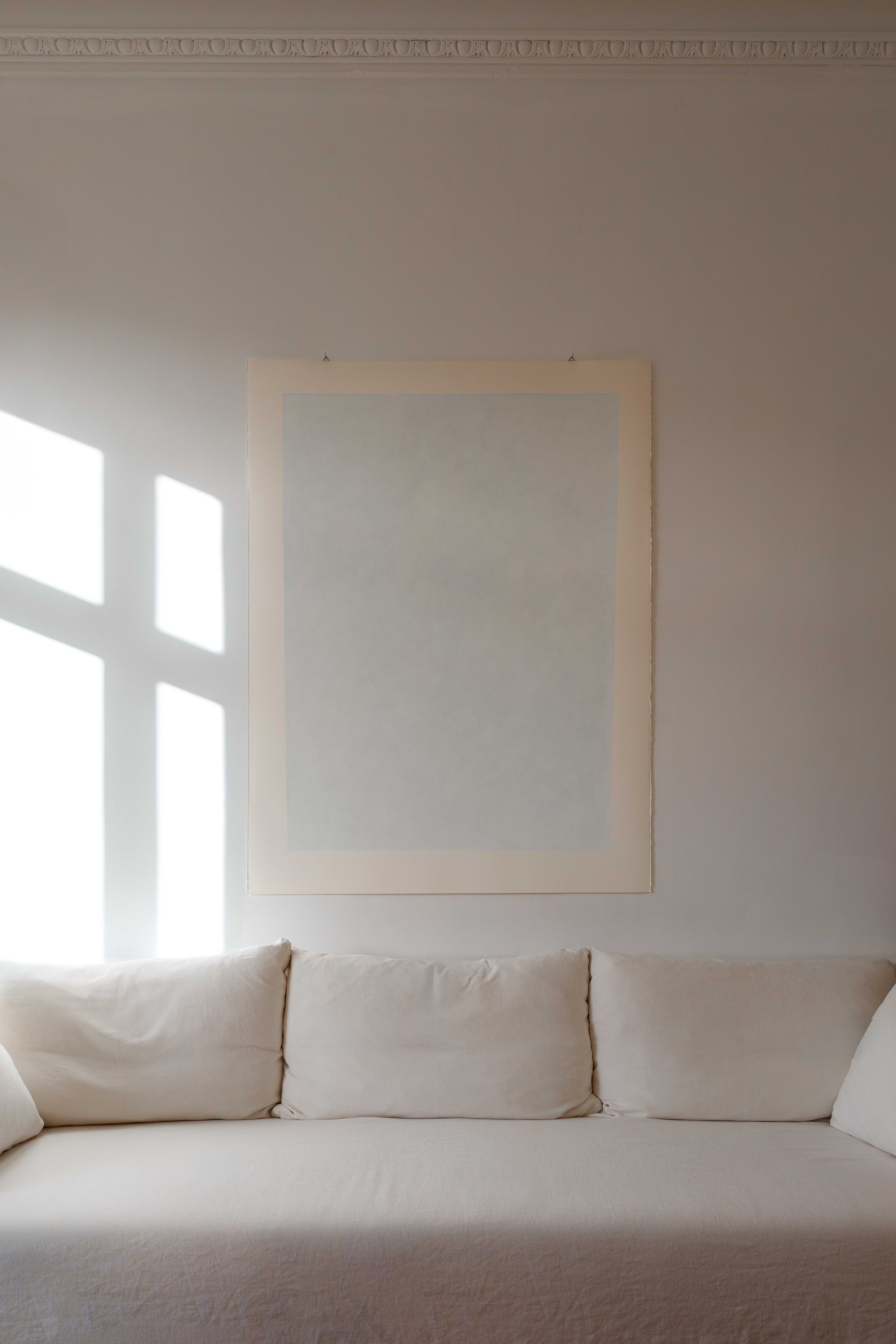 Minimalist living room with a white sofa, large abstract artwork on the wall, and sunlight casting a window-shaped shadow.