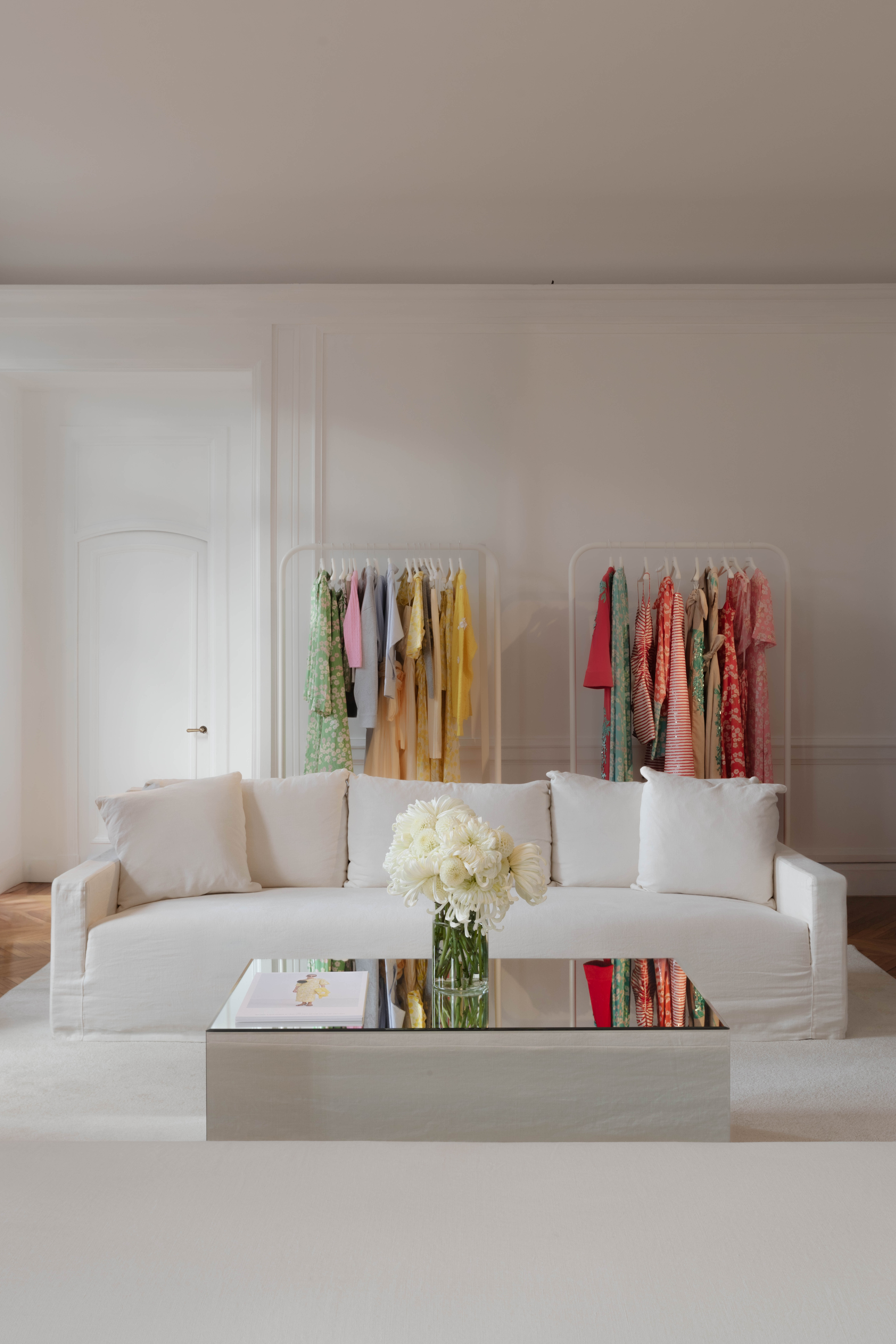 Minimalist living room with a white sofa, mirrored coffee table, and clothing racks displaying colorful garments in the background.