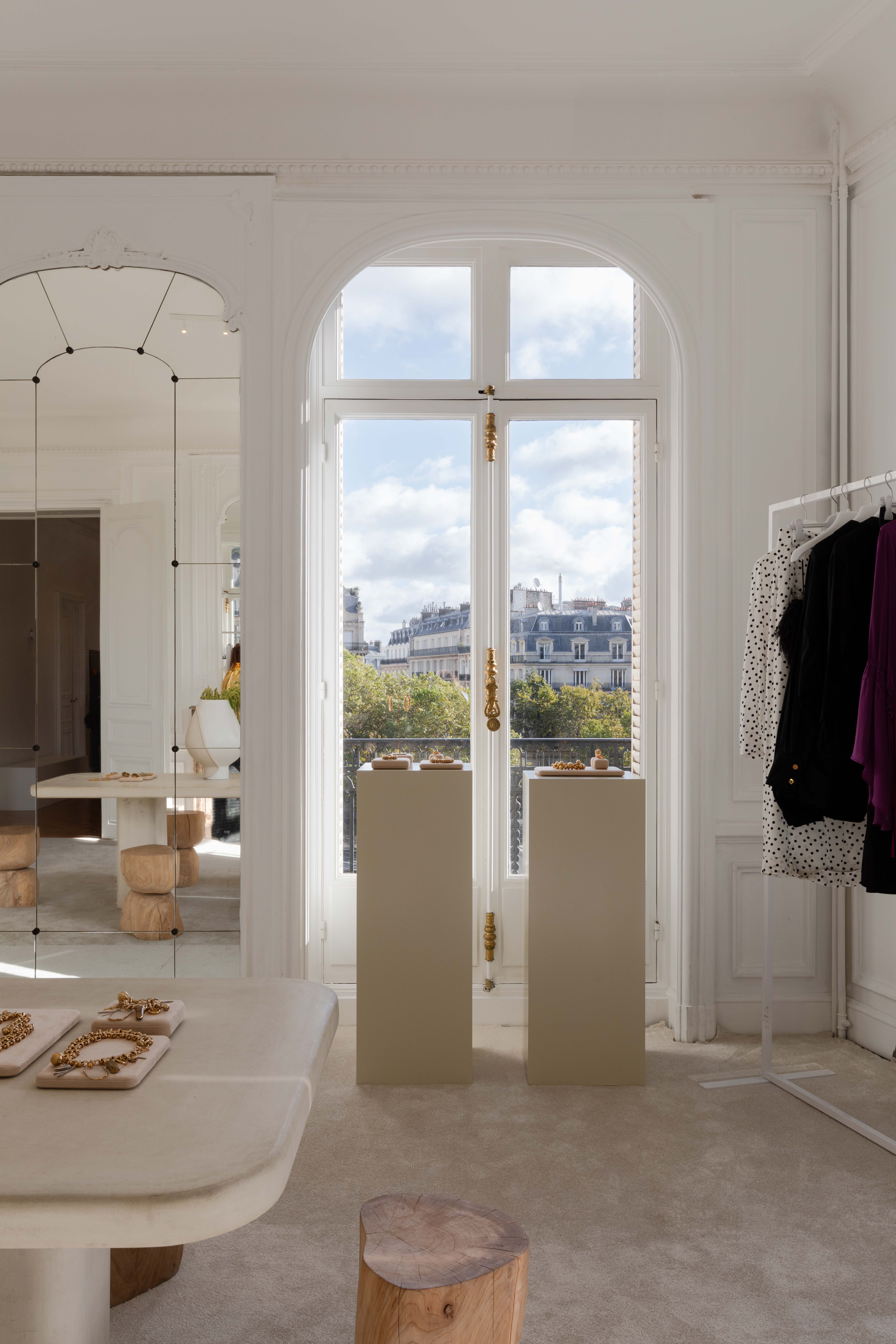 Elegant room with large arched window, displaying clothing and accessories. Bright natural light and a view of city buildings outside.
