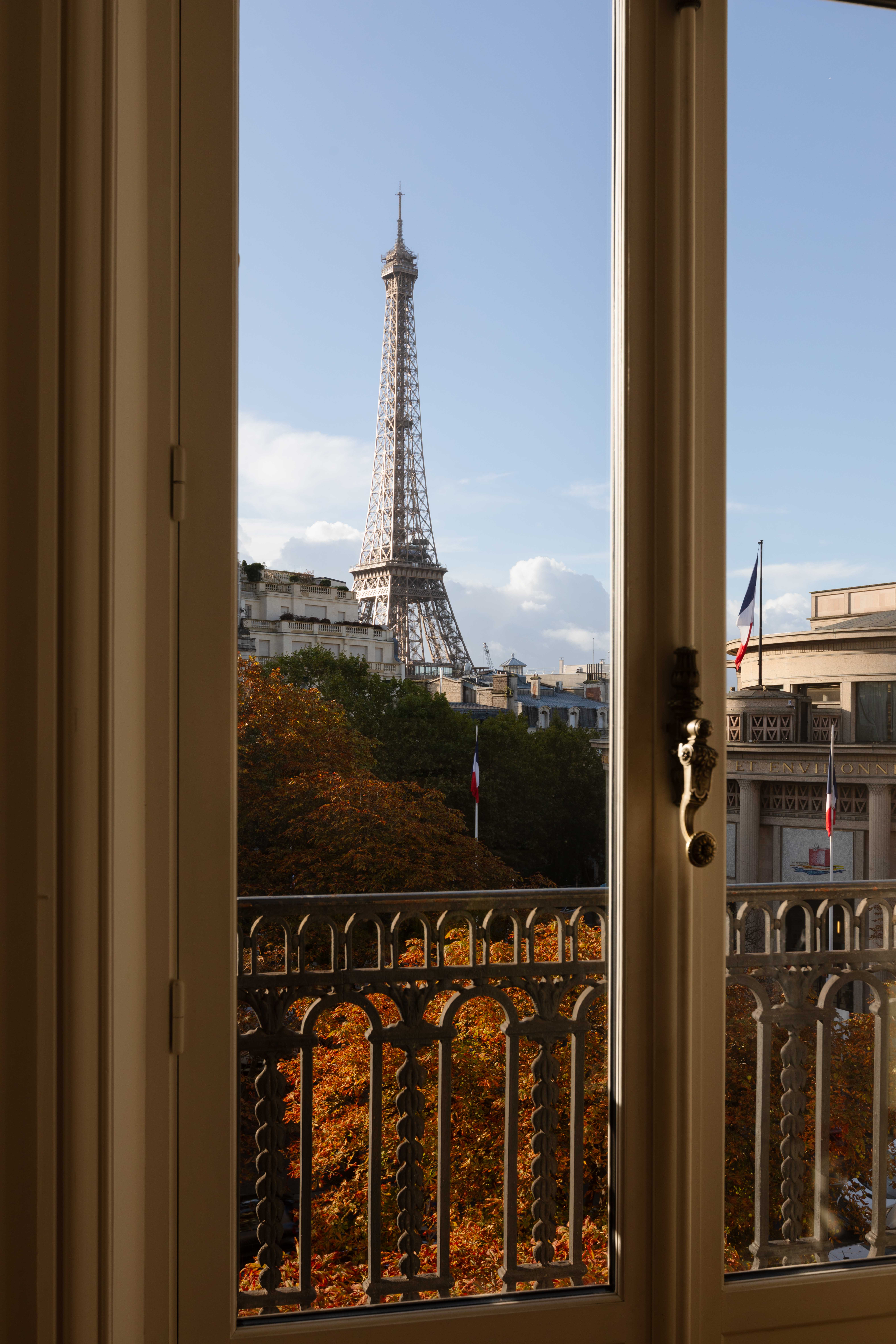 View of the Eiffel Tower through an open window, with autumn trees and Parisian buildings in the foreground under a clear blue sky.