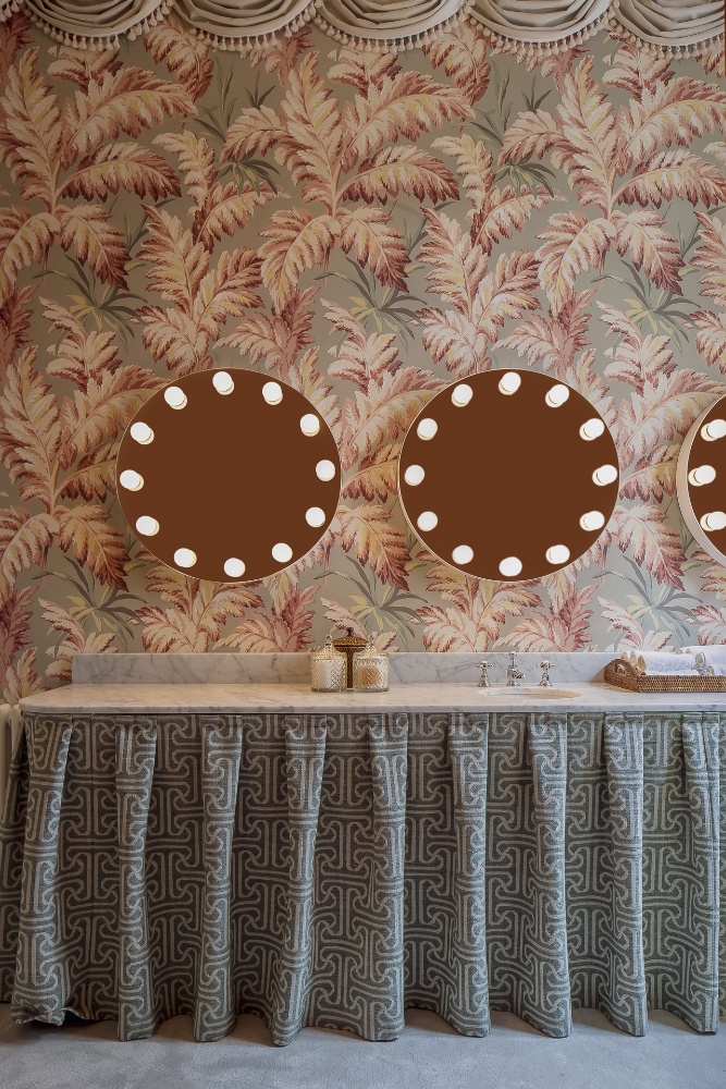 Elegant powder room with tropical wallpaper, round mirrors with lights, marble countertop, and fabric-draped vanity in The Nine Hotel.