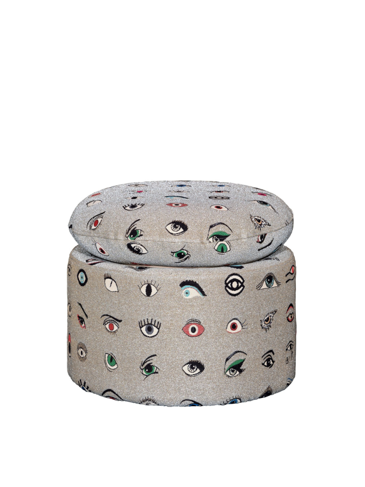 Olympia pouf in round shape, upholstered in a silver-grey fabric patterned with embroidered multicolored eyes in various styles and expressions, topped with a matching round cushion.