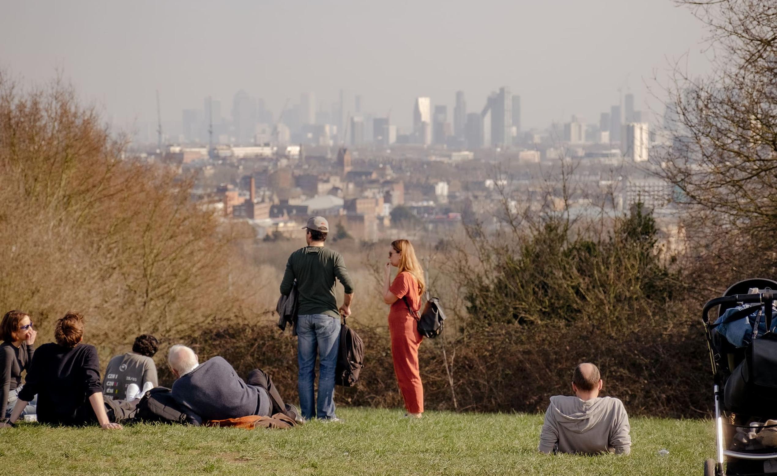 People relaxing on a grassy hill overlooking a city skyline in the distance, with trees and a stroller nearby.