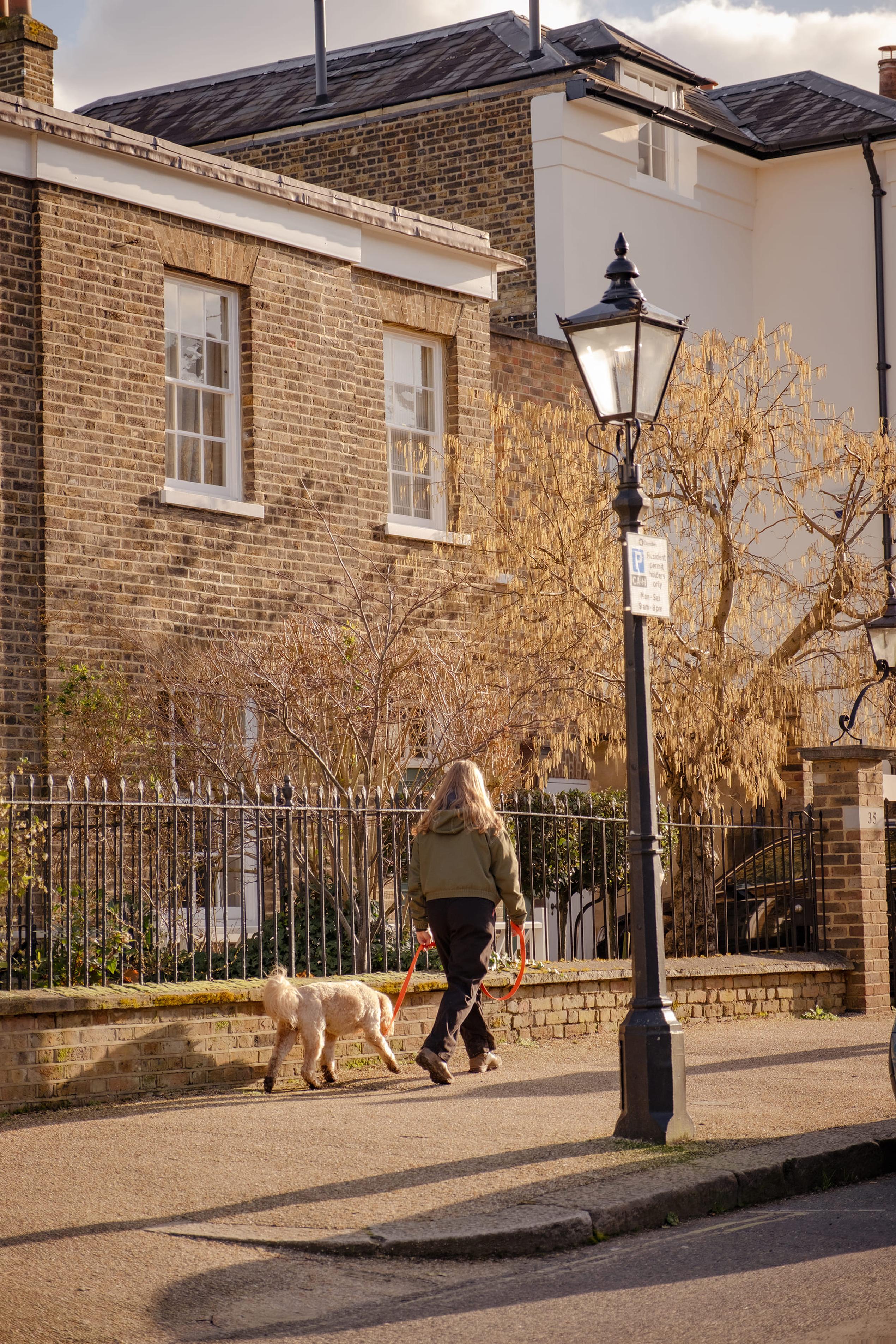 A person walks a dog on a leash down a sunny street with brick buildings and a vintage-style streetlamp.