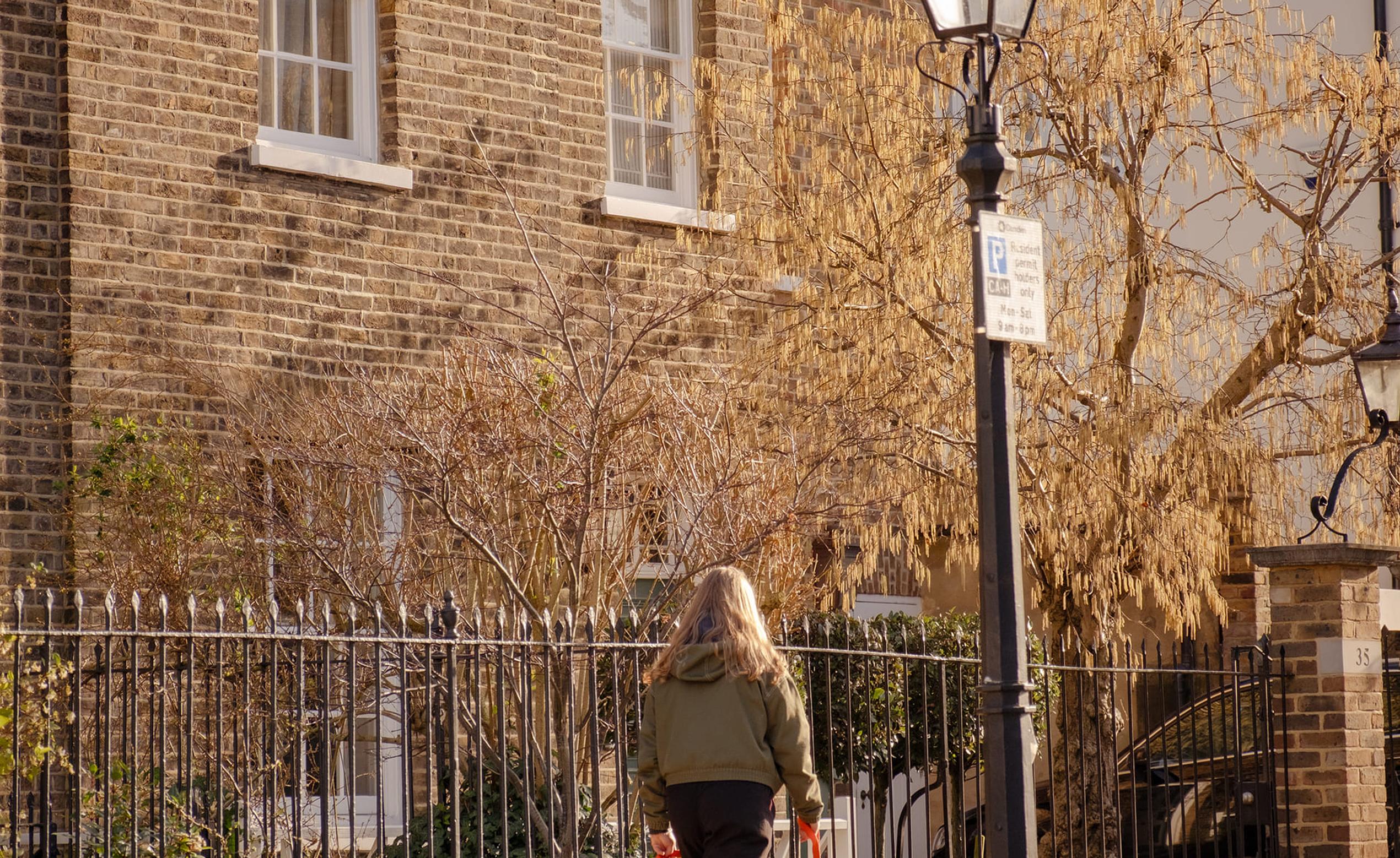 A person walks a dog on a leash down a sunny street with brick buildings and a vintage-style streetlamp.