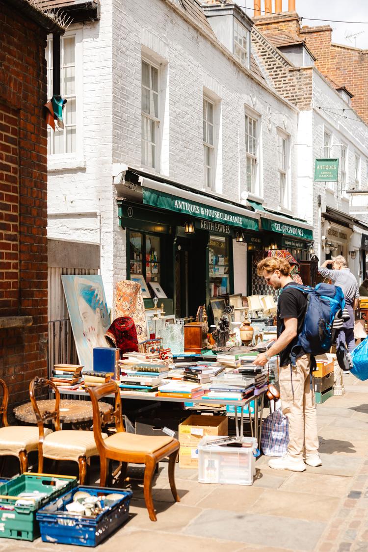 A man browses books and antiques outside a shop with green awnings on a sunny street. Various items and furniture are displayed around him.