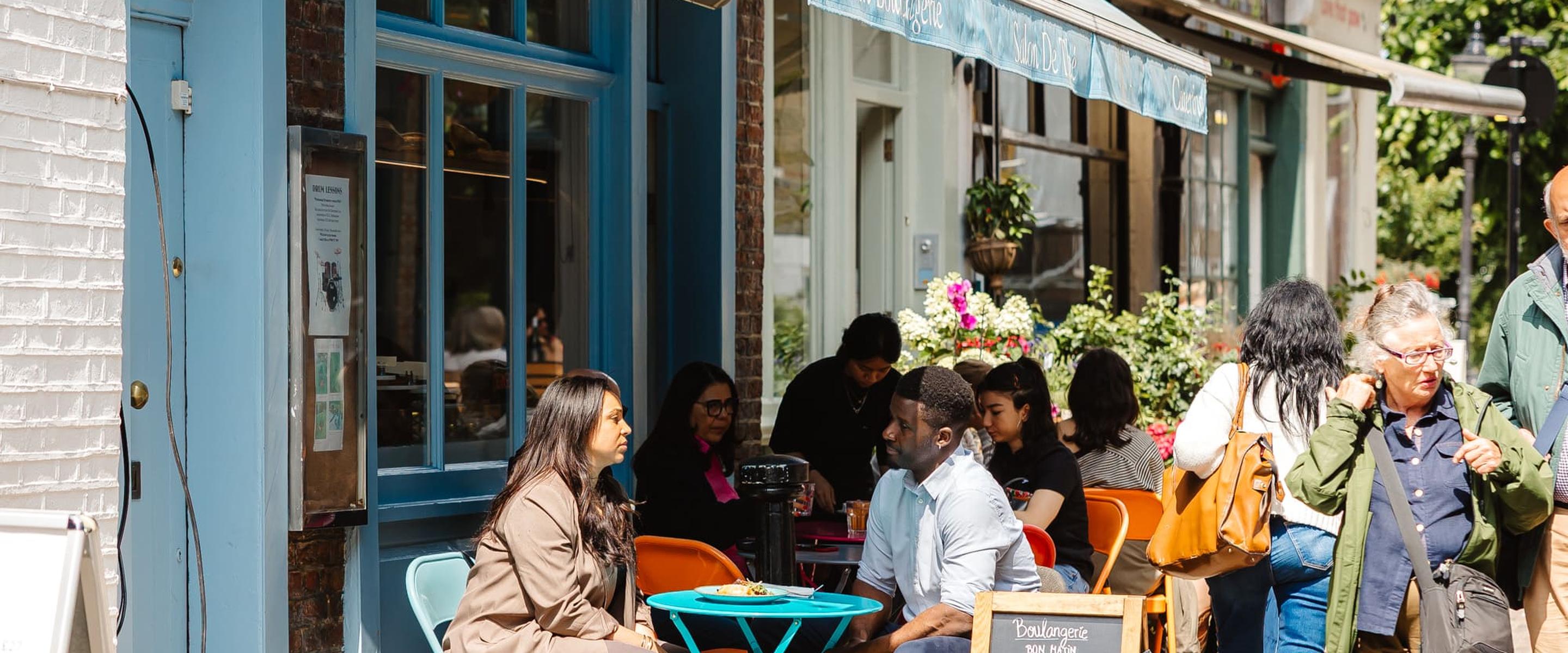 People sitting outside a blue boulangerie, enjoying a sunny day at turquoise tables with a chalkboard menu by the entrance.
