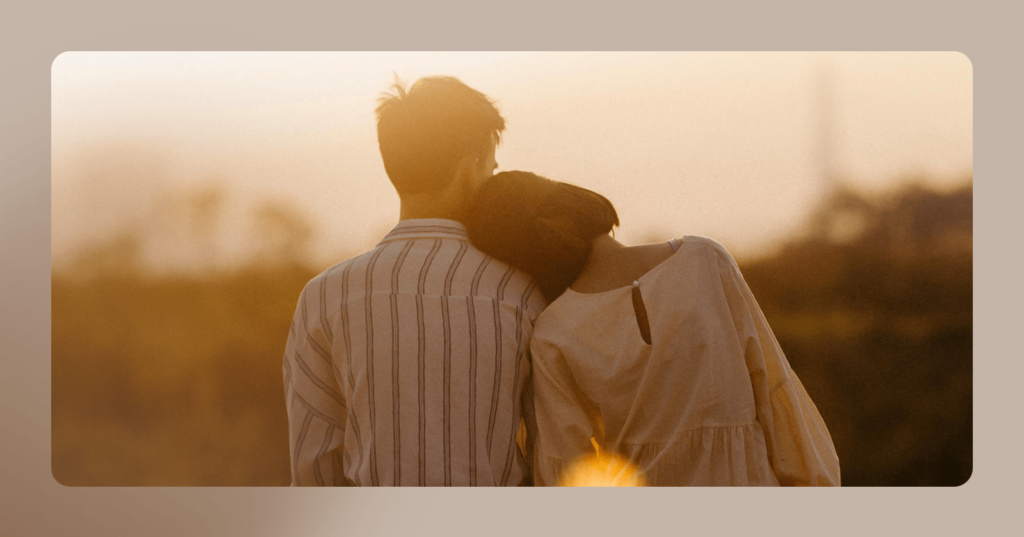 Image of couple looking against a sunset skyline, one is leaning on the other's shoulder. The image is framed in tan.