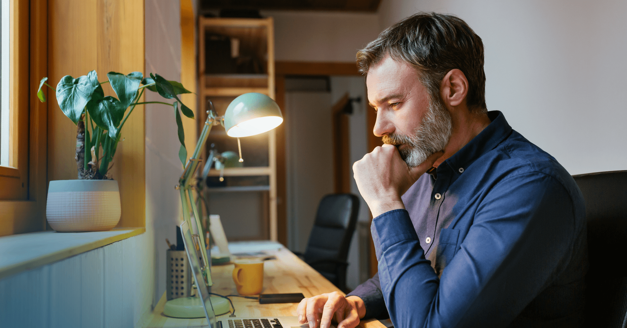 Man sits in a black leather chair staring down intently at the laptop next to a bright lamp in front of him. His left hand is on his chin as he scrolls with his right hand. He is wearing a dark blue button up shirt. The background is a white wall.