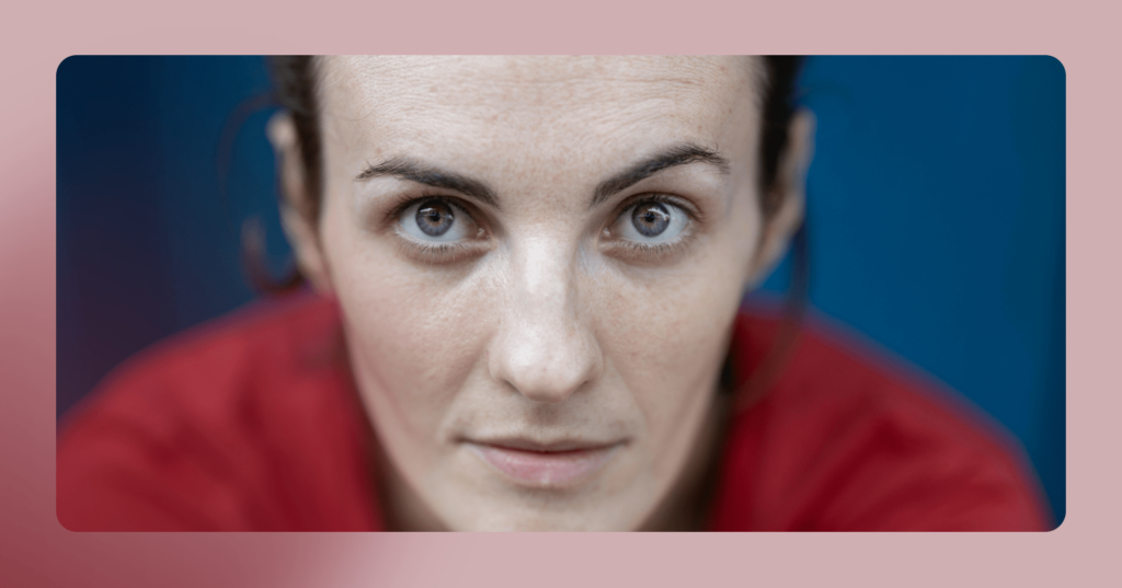 Woman sits staring straight into the camera. It is zoomed in on her face down to her shoulders. She is wearing a red T-shirt and the background is blue. There is a pink border around the image.