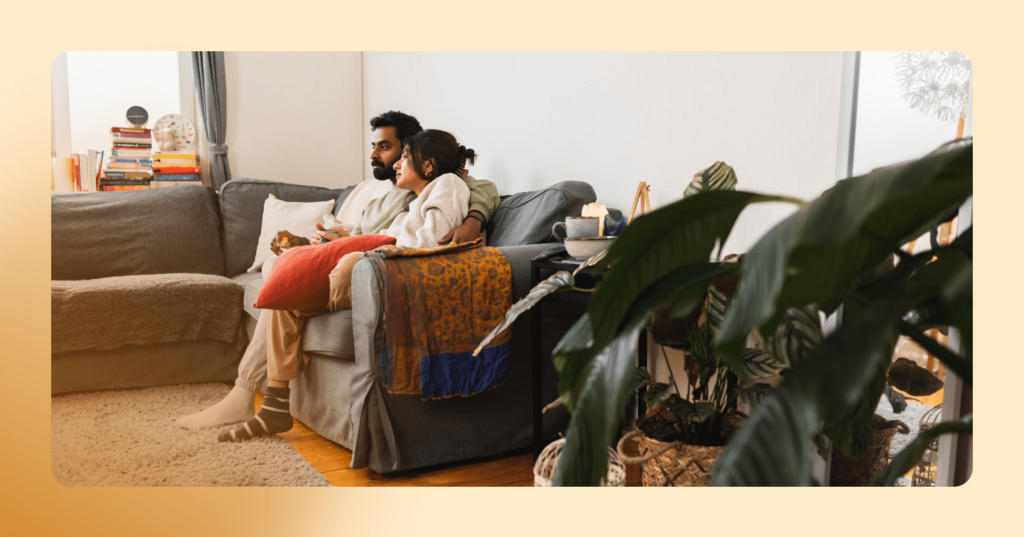 A man and woman are sitting on a couch together. The man looks to be upset. The photo shows a living room with plants and stacks of books throughout. The image is framed in orange.