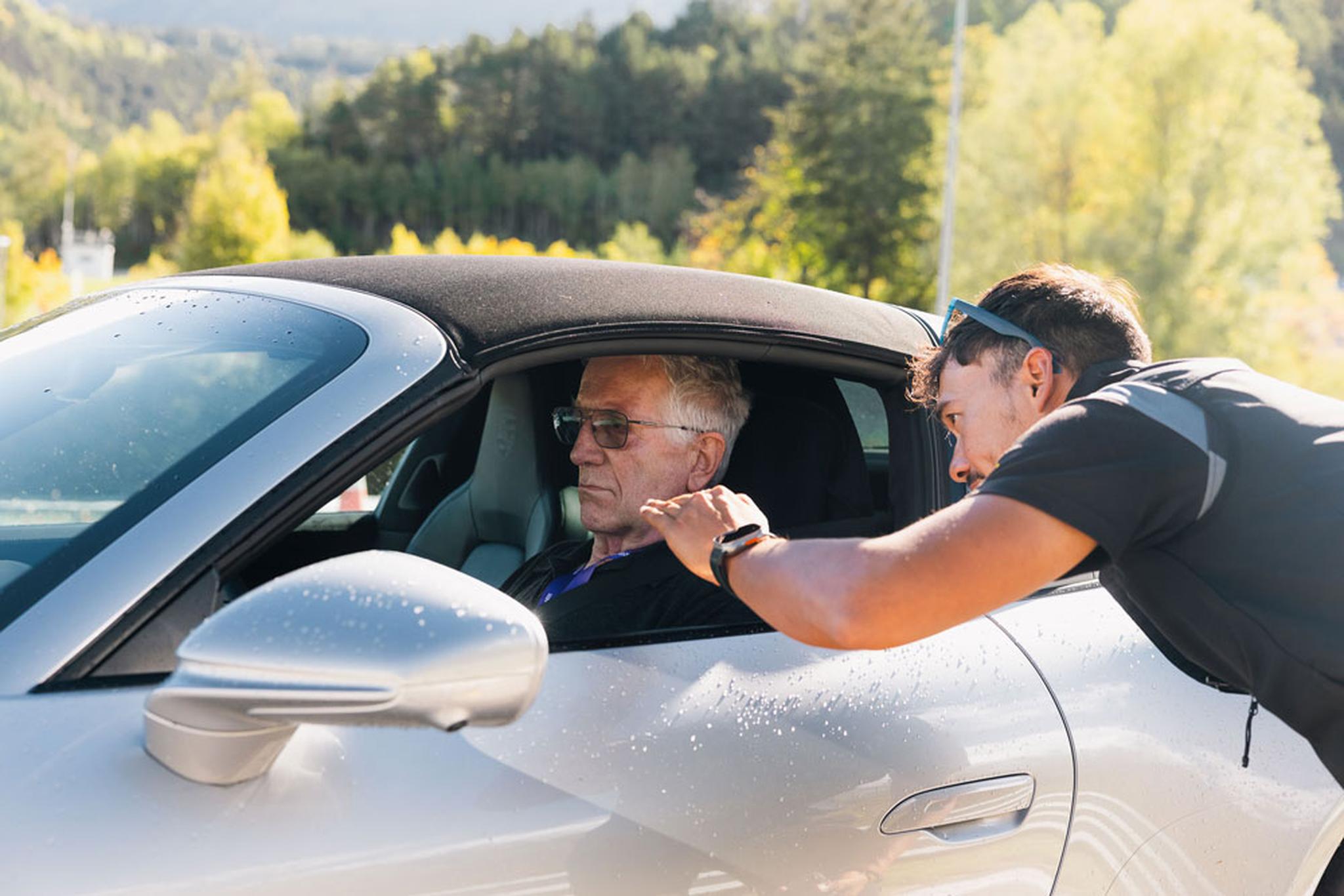 Ein Mann mit Sonnenbrille beugt sich vor, um mit einem älteren Mann zu sprechen, der in einem Auto sitzt. Das Auto ist silberfarben und steht an einem sonnigen Tag im Freien.