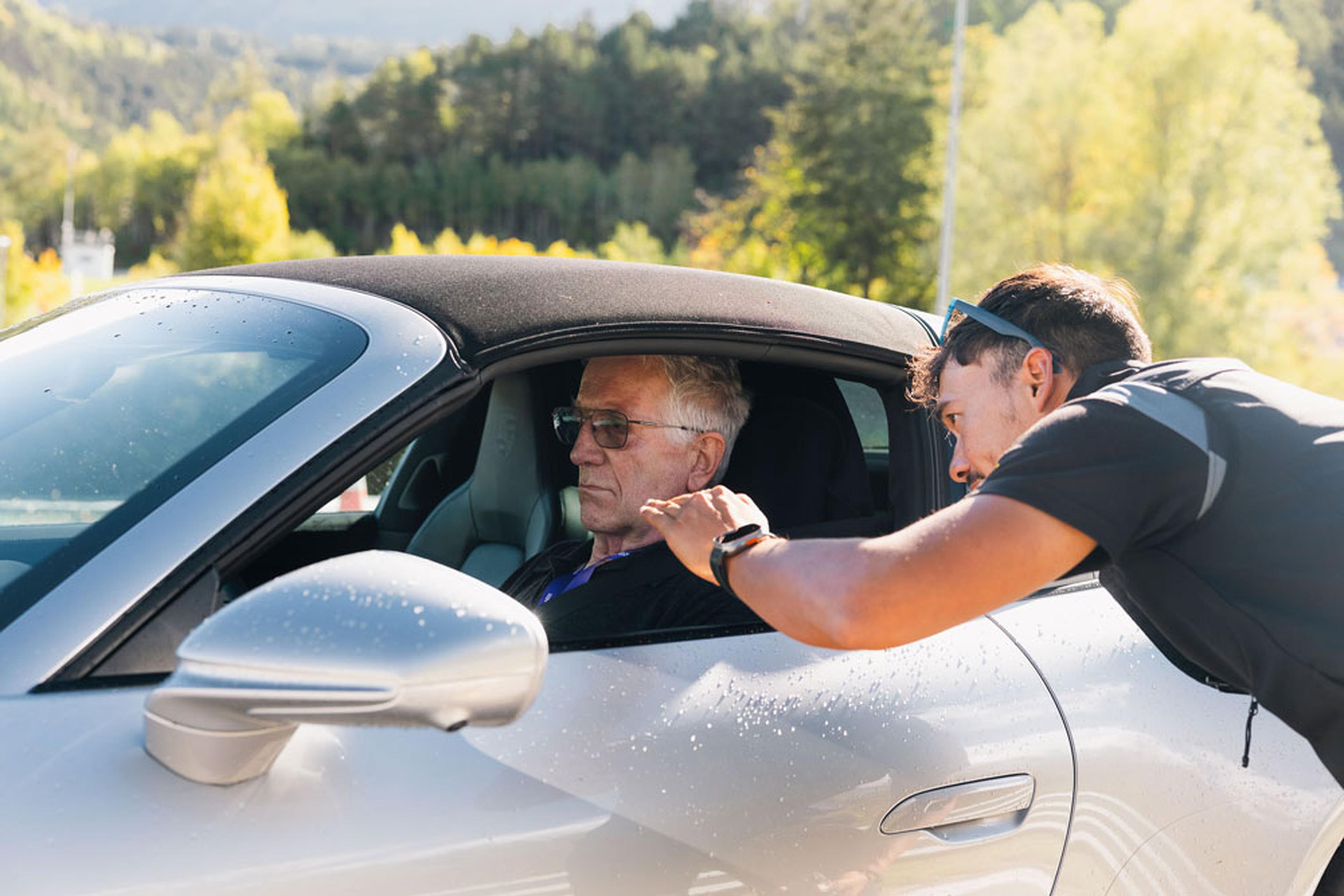 Ein Mann mit Sonnenbrille beugt sich vor, um mit einem älteren Mann zu sprechen, der in einem Auto sitzt. Das Auto ist silberfarben und steht an einem sonnigen Tag im Freien.