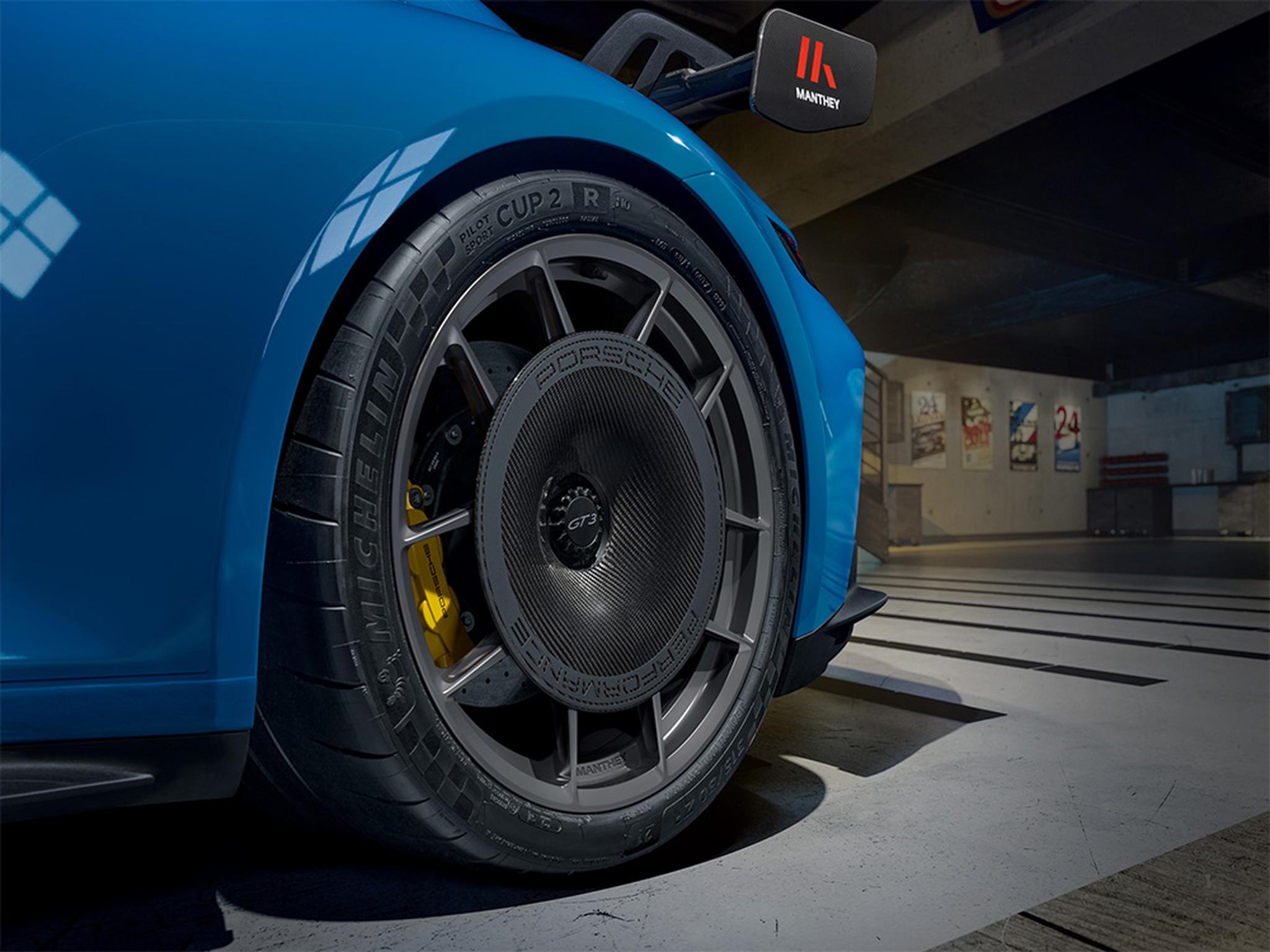 Close-up of a Manthey lightweight wheel fitted to a blue Porsche 911, featuring a center-mounted aerodynamic wheel cover, Michelin performance tire, and high-performance brake system.