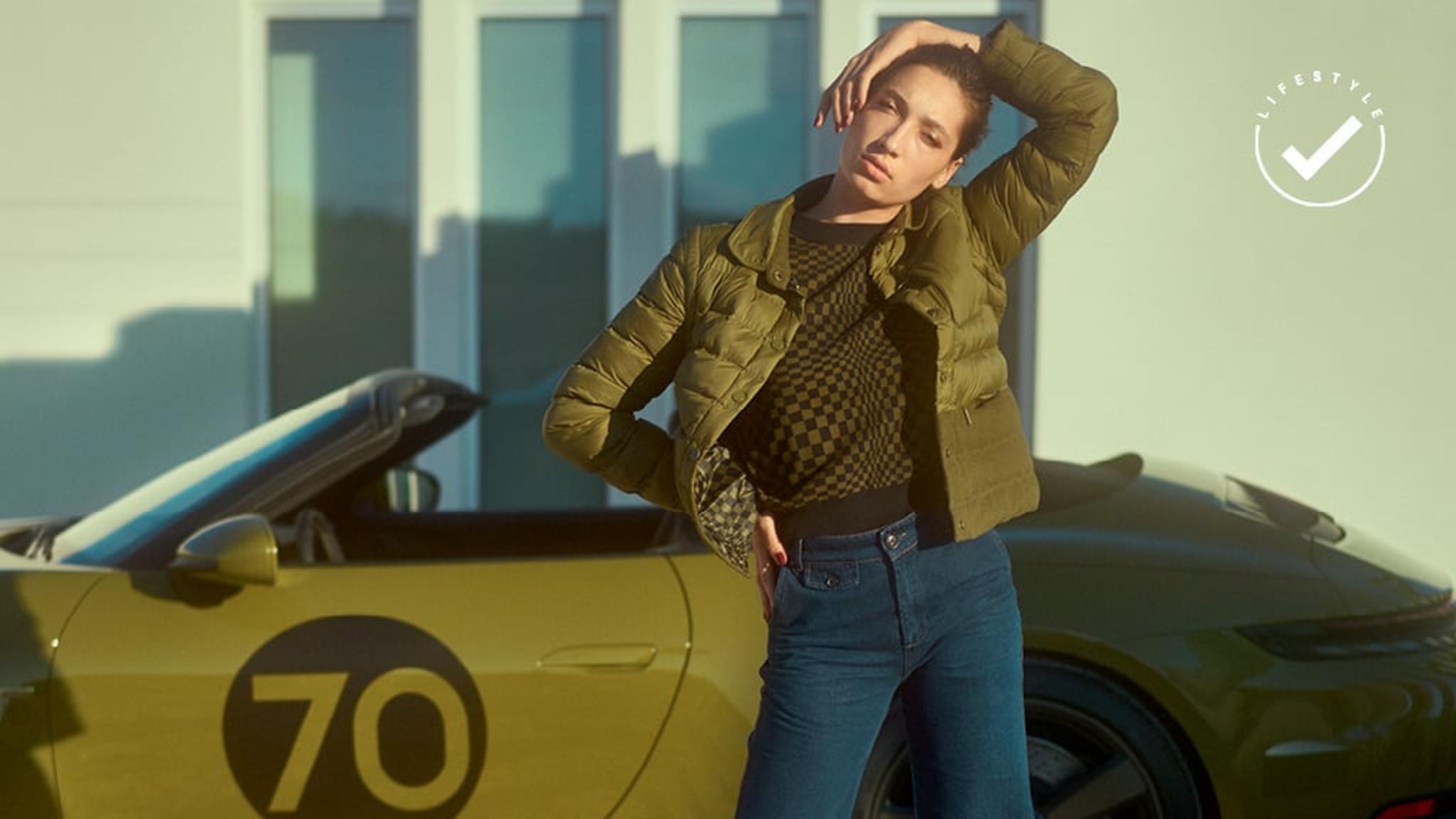 Mujer en chaqueta acolchada verde y jeans, posando casualmente frente a un llamativo Porsche 911 Targa Heritage Design con el número 70