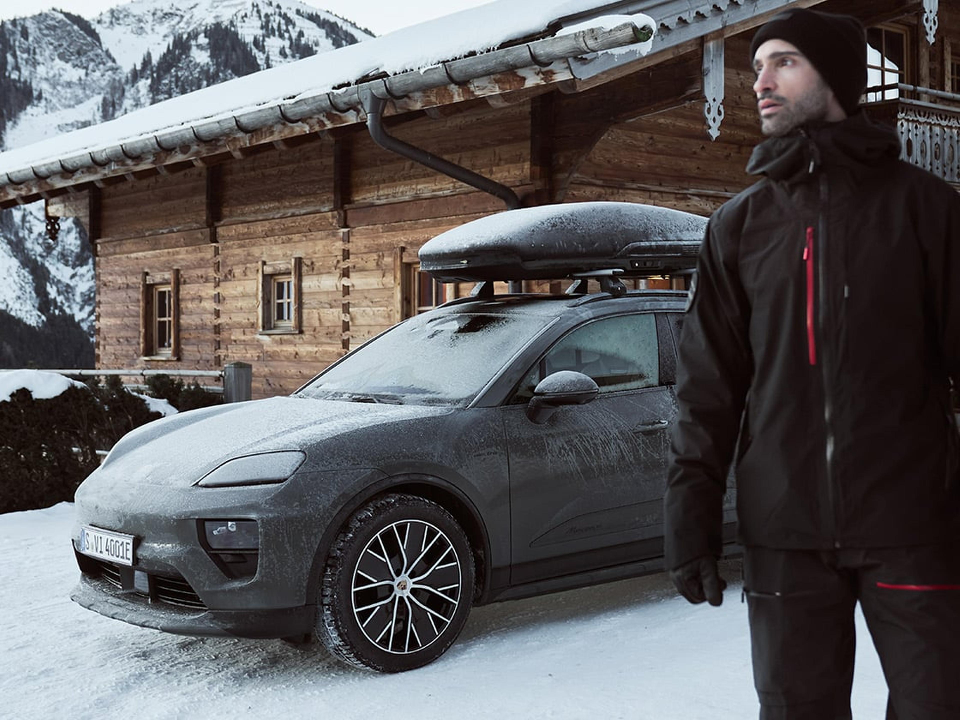 Porsche mit Dachbox steht vor einer Hütte im Schnee und ein Mann steht vor dem Auto.