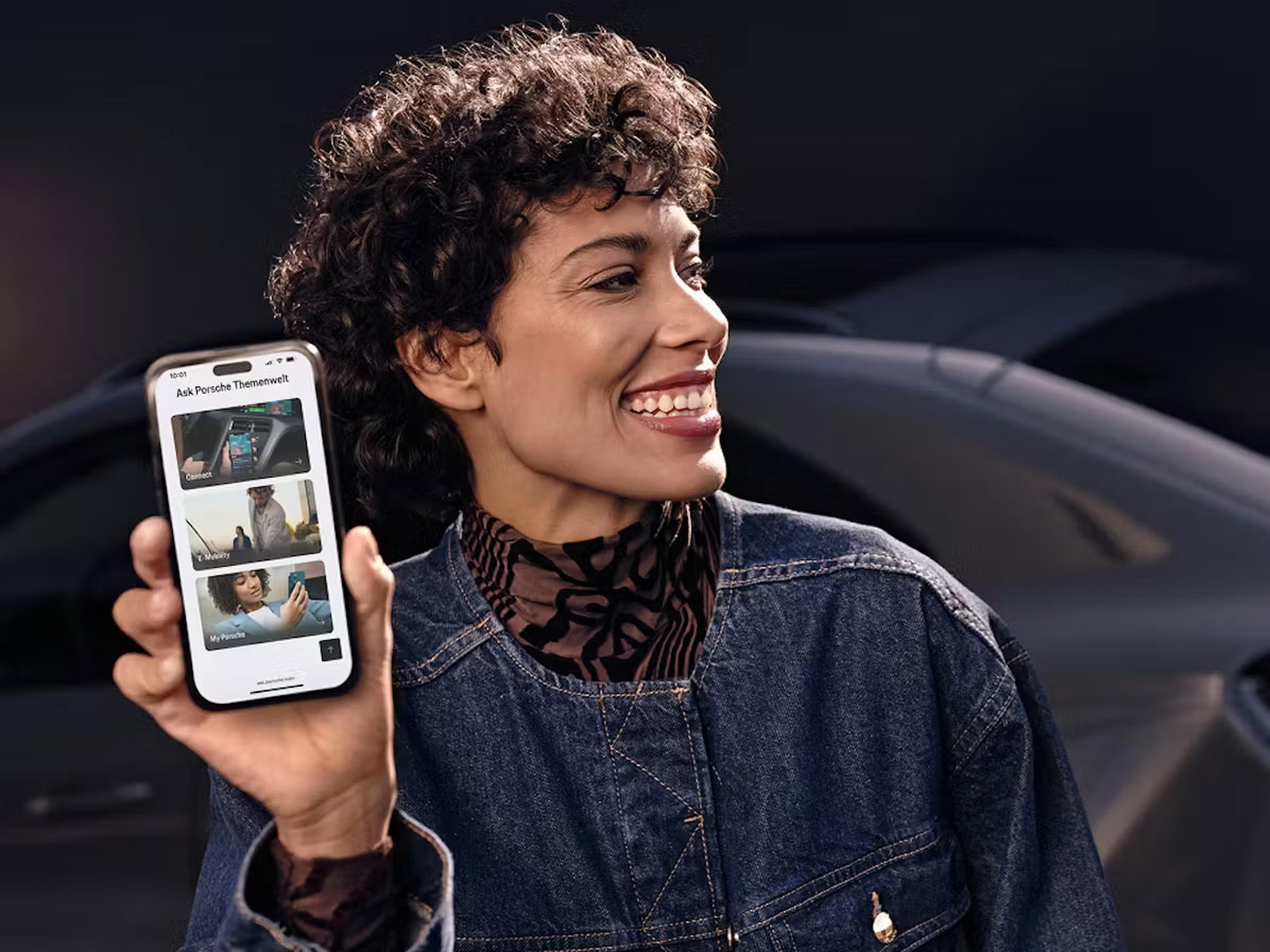 Eine lächelnde Person steht vor einem Auto und hält ein Smartphone, auf dem Bilder zu sehen sind.