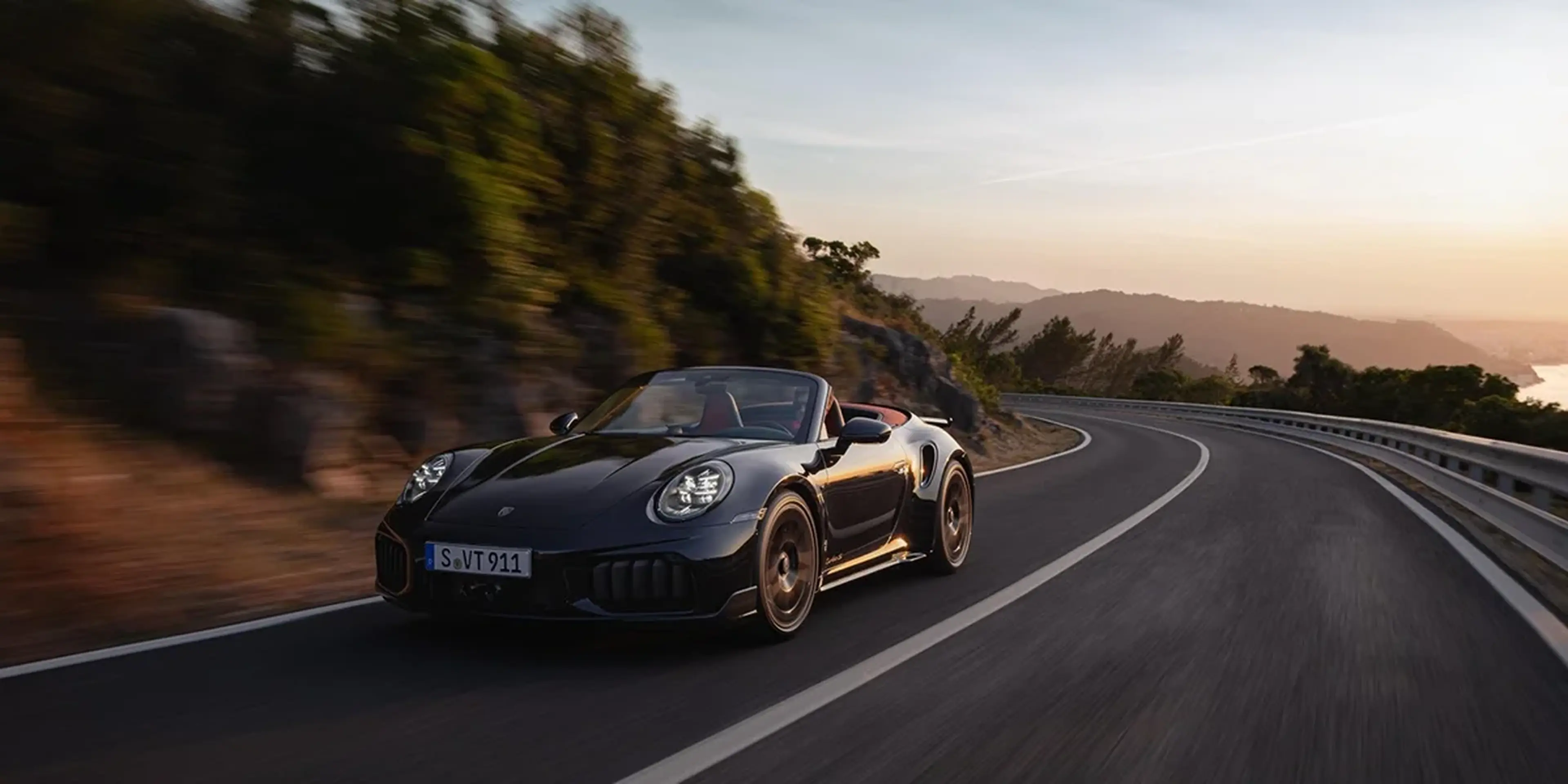 Porsche 911 Turbo S Cabriolet driving along a coastal road at sunrise, dynamic front angle.
