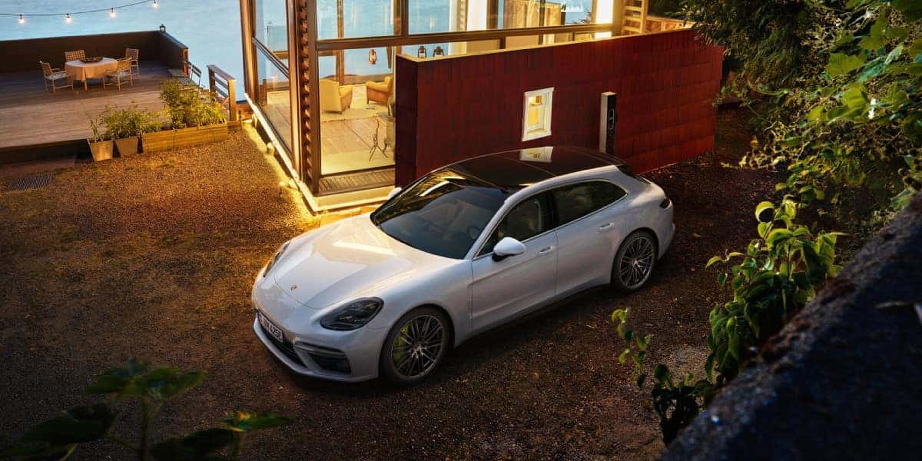 A sleek white car parked next to a modern glass house at dusk, with warm lighting and outdoor seating visible in the background.