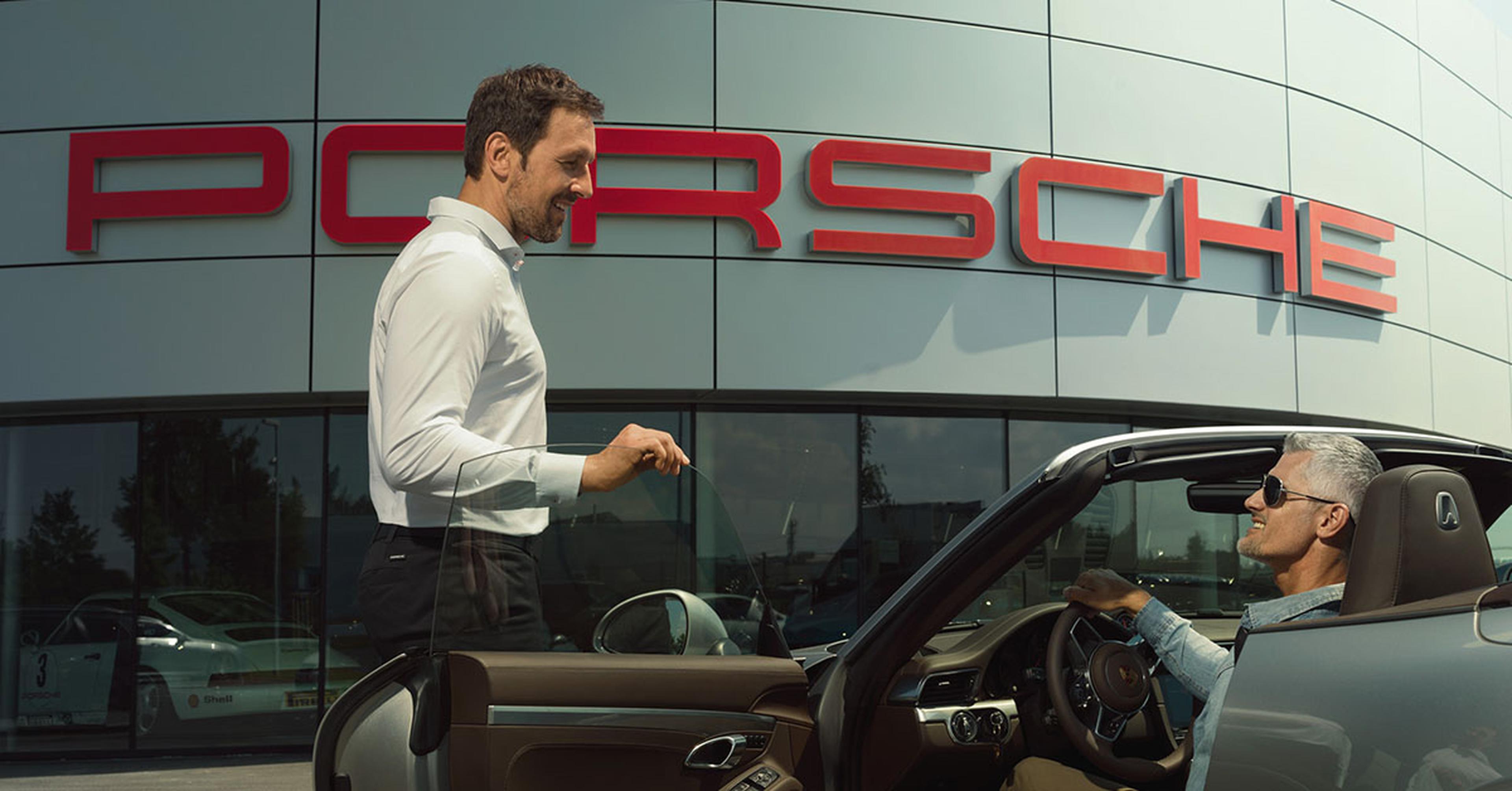 Man in white shirt standing beside a man in sunglasses sitting in a convertible Porsche, in front of a Porsche dealership building.