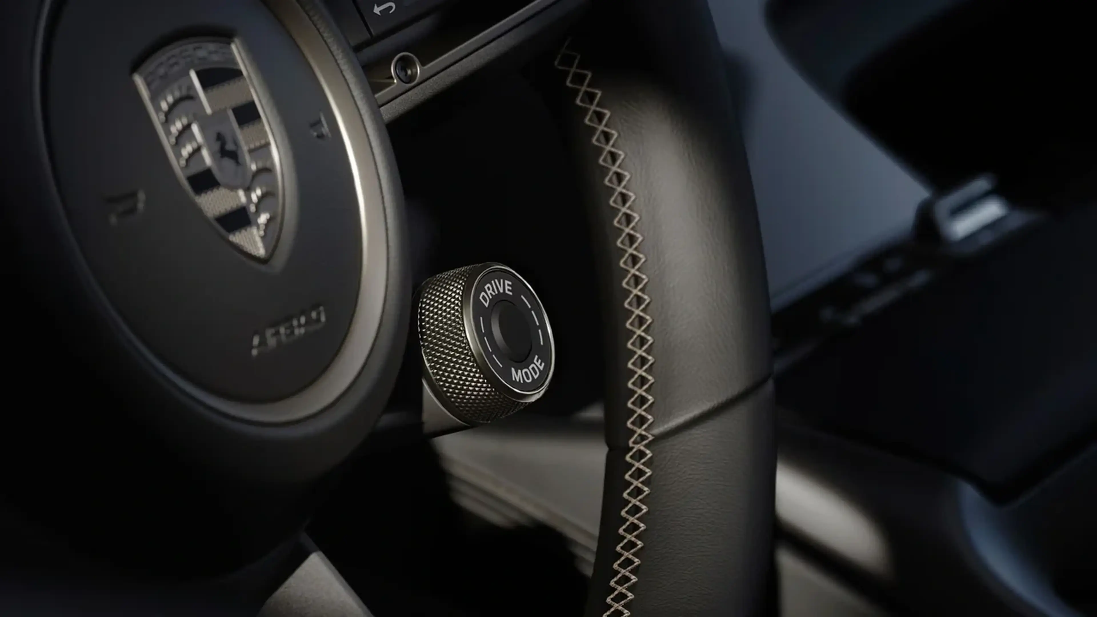 Close-up of a Porsche steering wheel with a mode selector dial, featuring intricate stitching and a Porsche crest visible on the wheel.