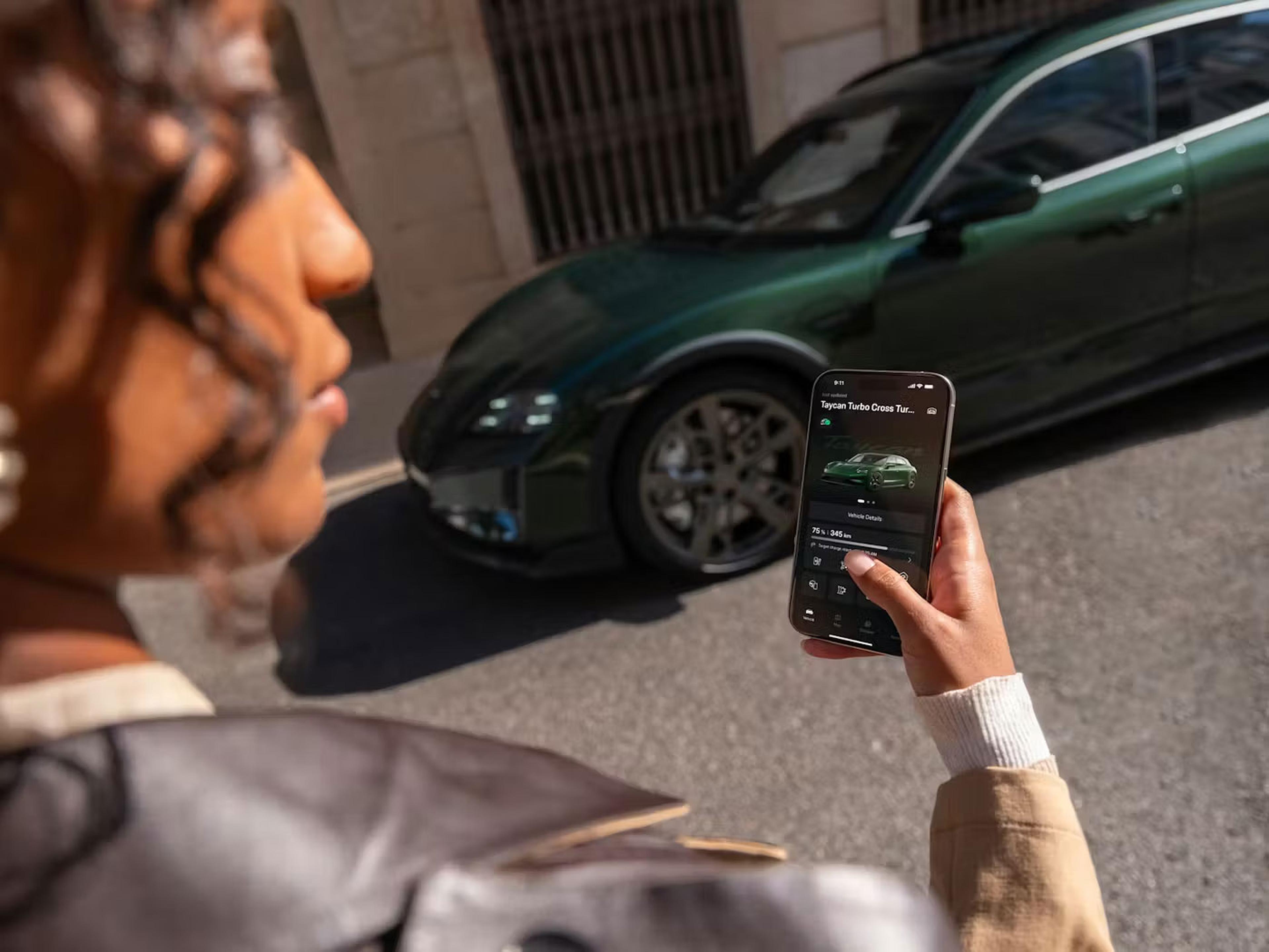 Eine Person hält ein Smartphone mit der Benutzeroberfläche einer Auto-App in der Hand und steht neben einem grünen Porsche, das an der Straße geparkt ist.