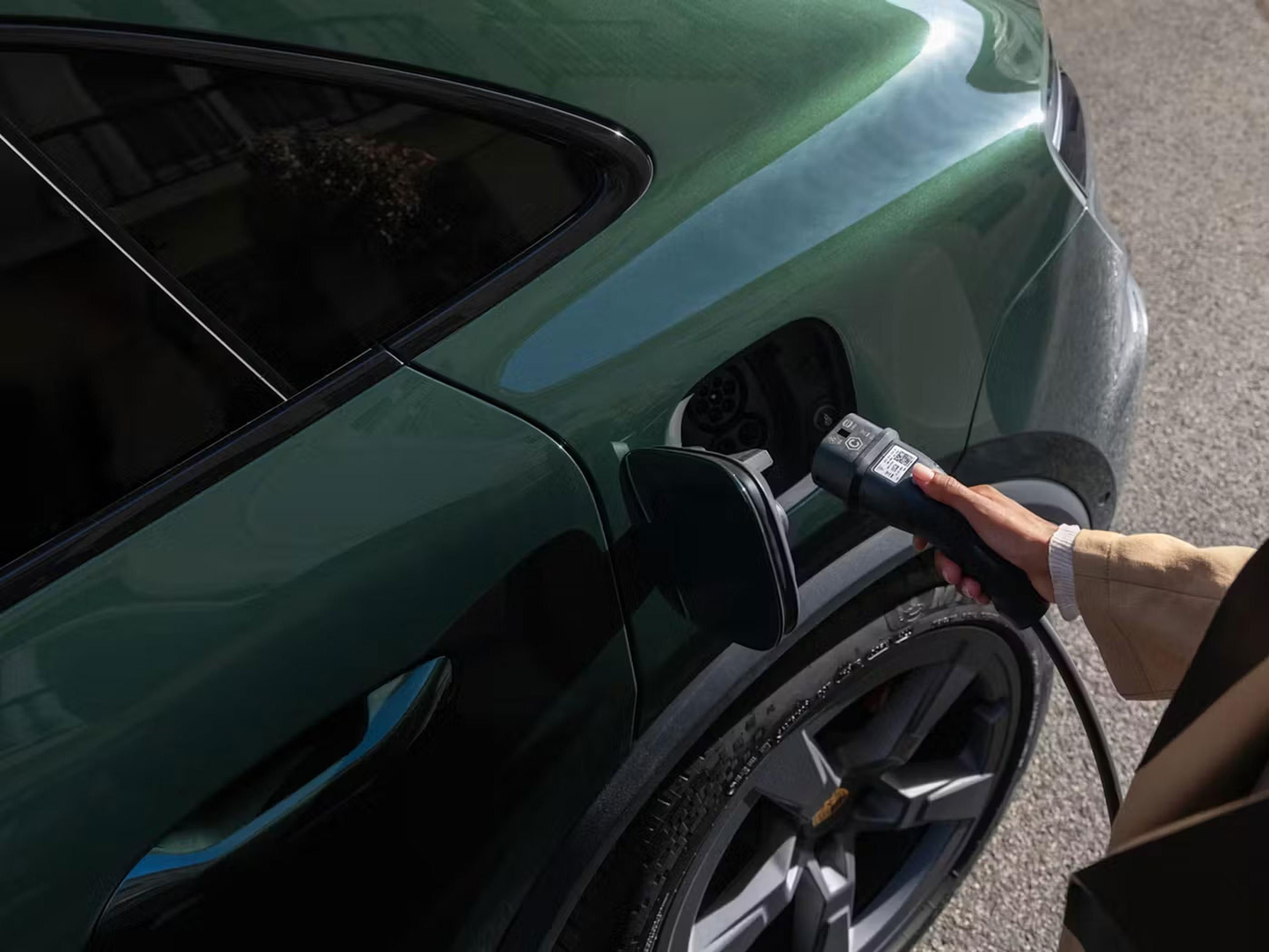 Eine Person steckt ein Ladekabel in ein dunkelgrünes Elektroauto, das an einem sonnigen Tag geparkt ist.