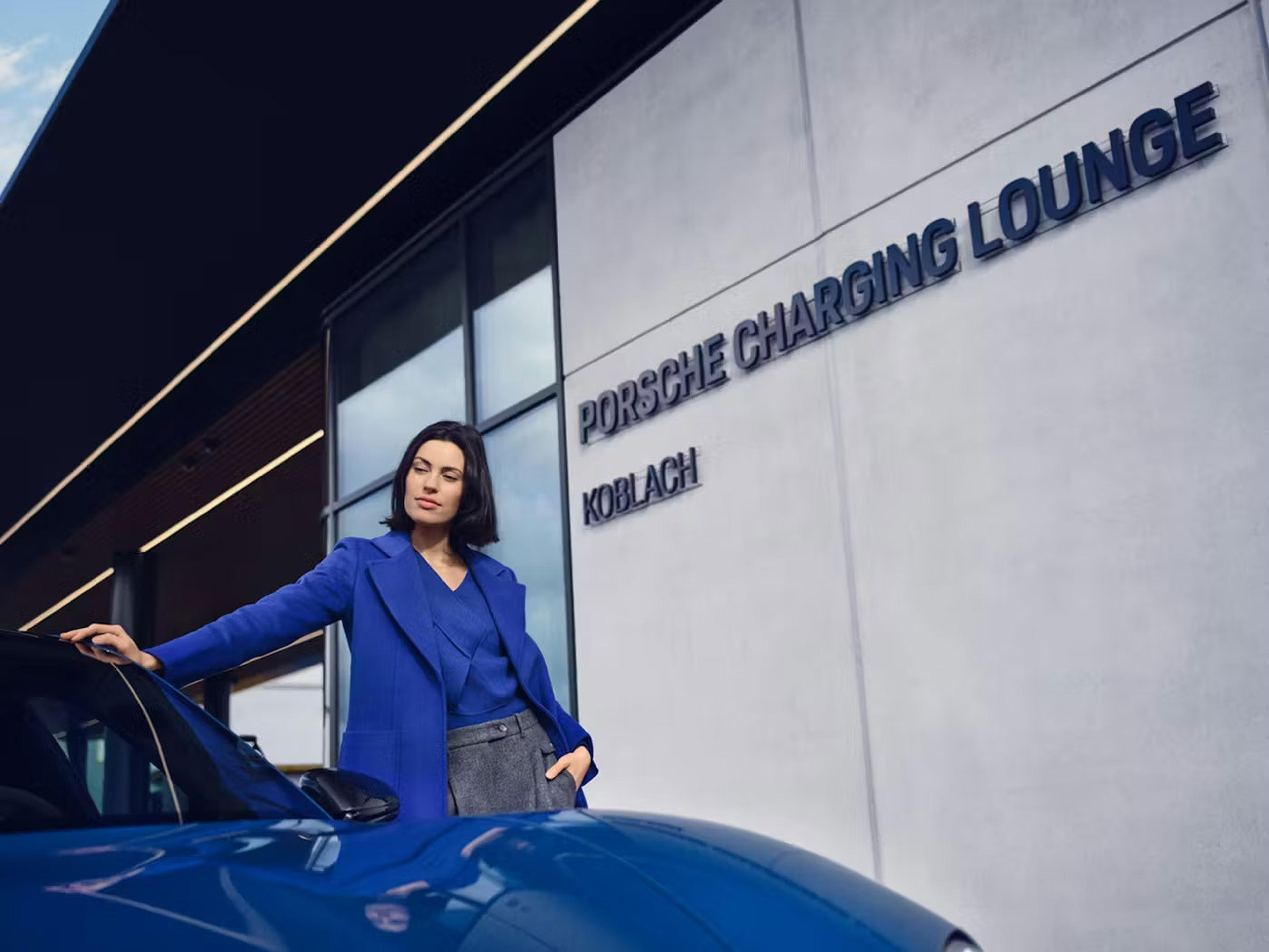Eine Frau in blauer Kleidung steht selbstbewusst neben einem blauen Auto; im Hintergrund ist am Gebäude das Schild „Porsche Charging Lounge Koblach“ zu sehen.