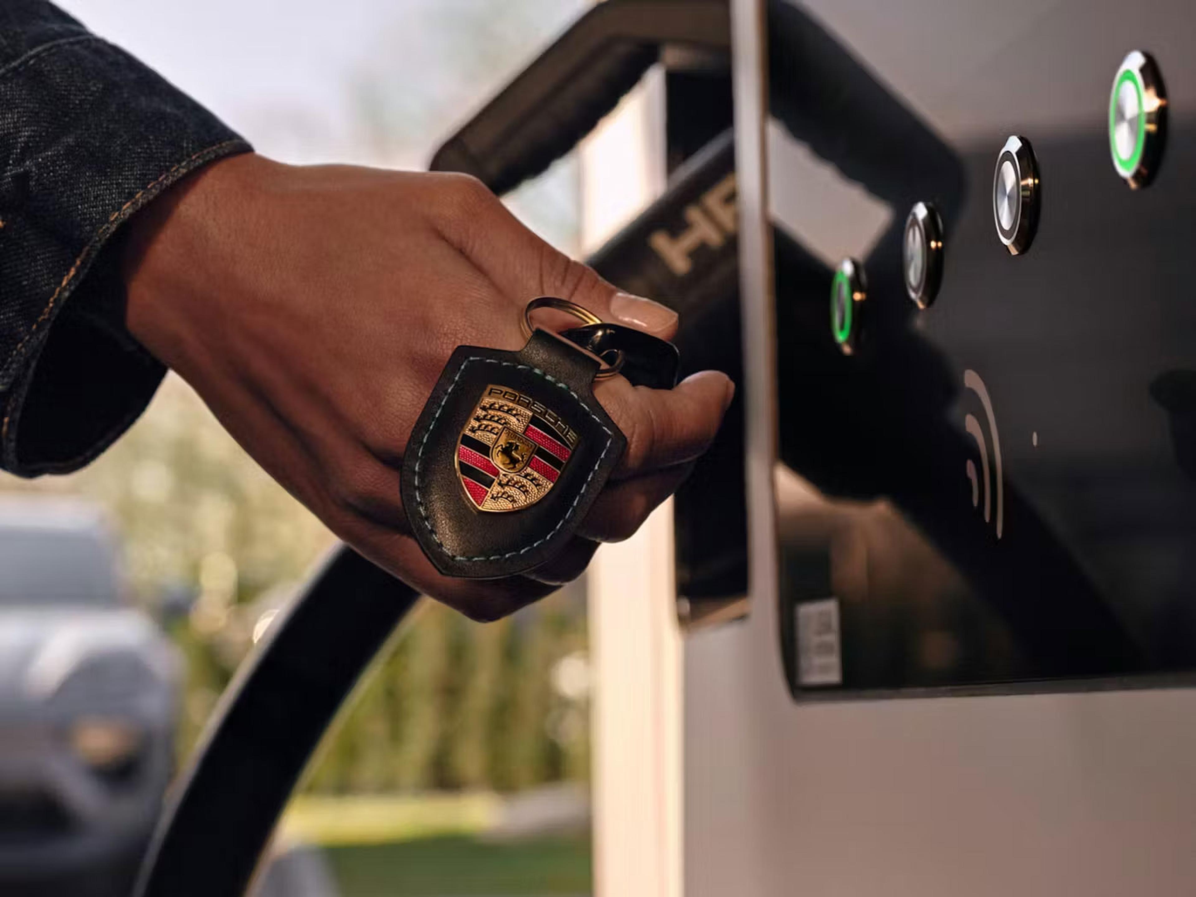 Eine Hand hält einen Schlüsselanhänger mit Porsche-Logo vor eine Ladestation mit grün beleuchteten Tasten; der Hintergrund ist unscharf.