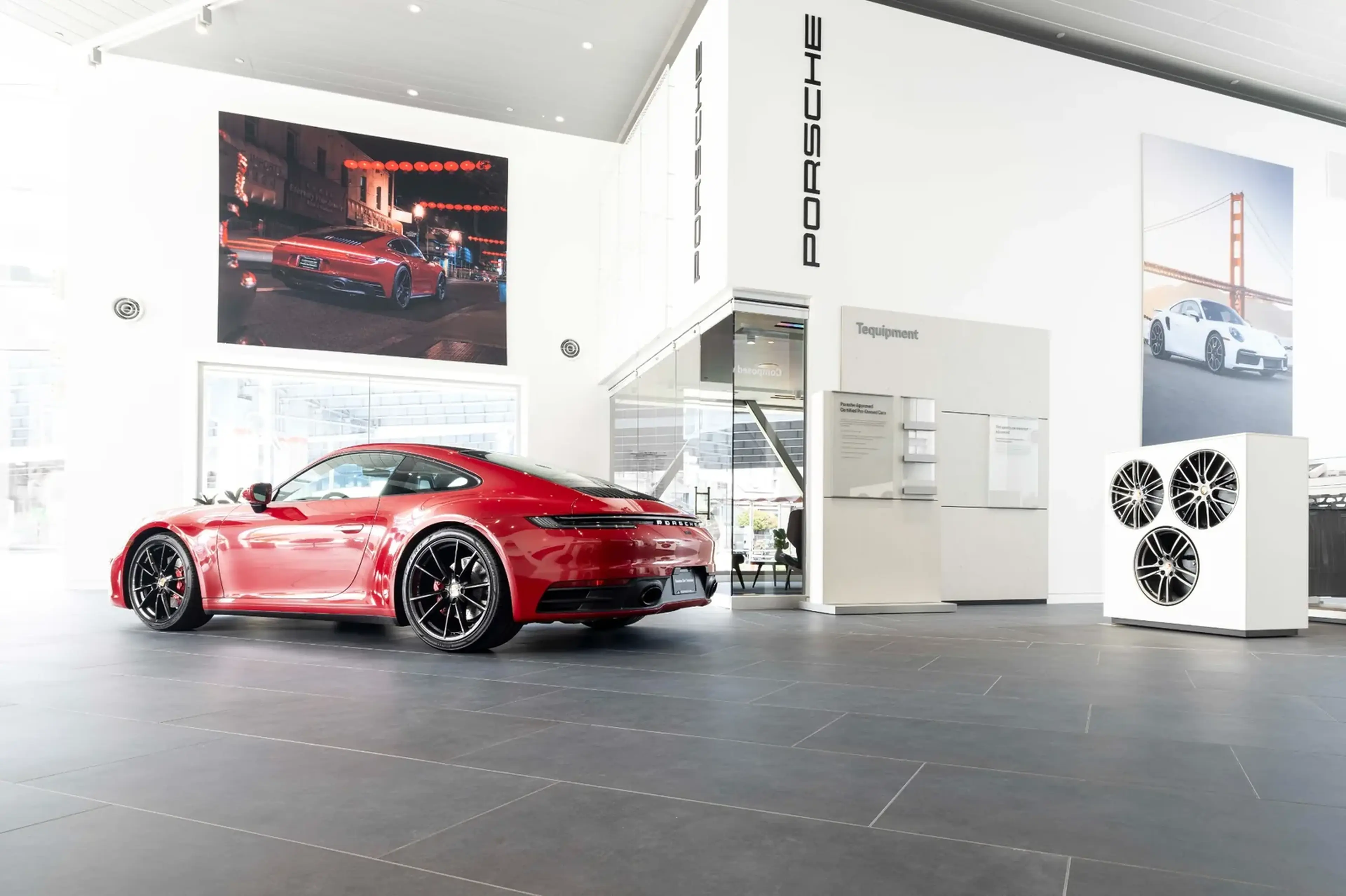 A red sports car is displayed in a bright, modern showroom with Porsche branding, large wall photos, and a display of car rims.