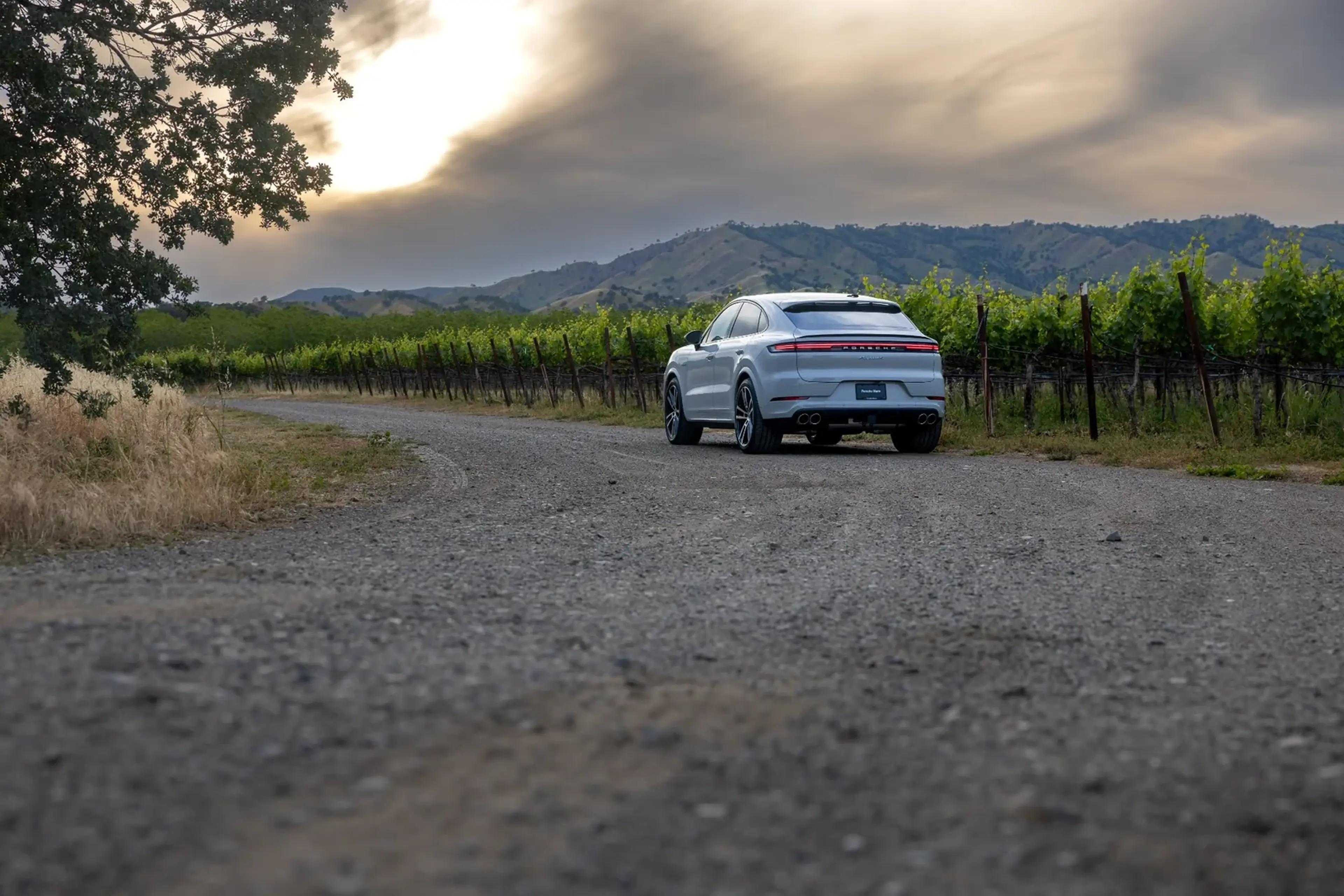 A white SUV on a gravel road beside a vineyard, with hills and a dramatic sky in the background.