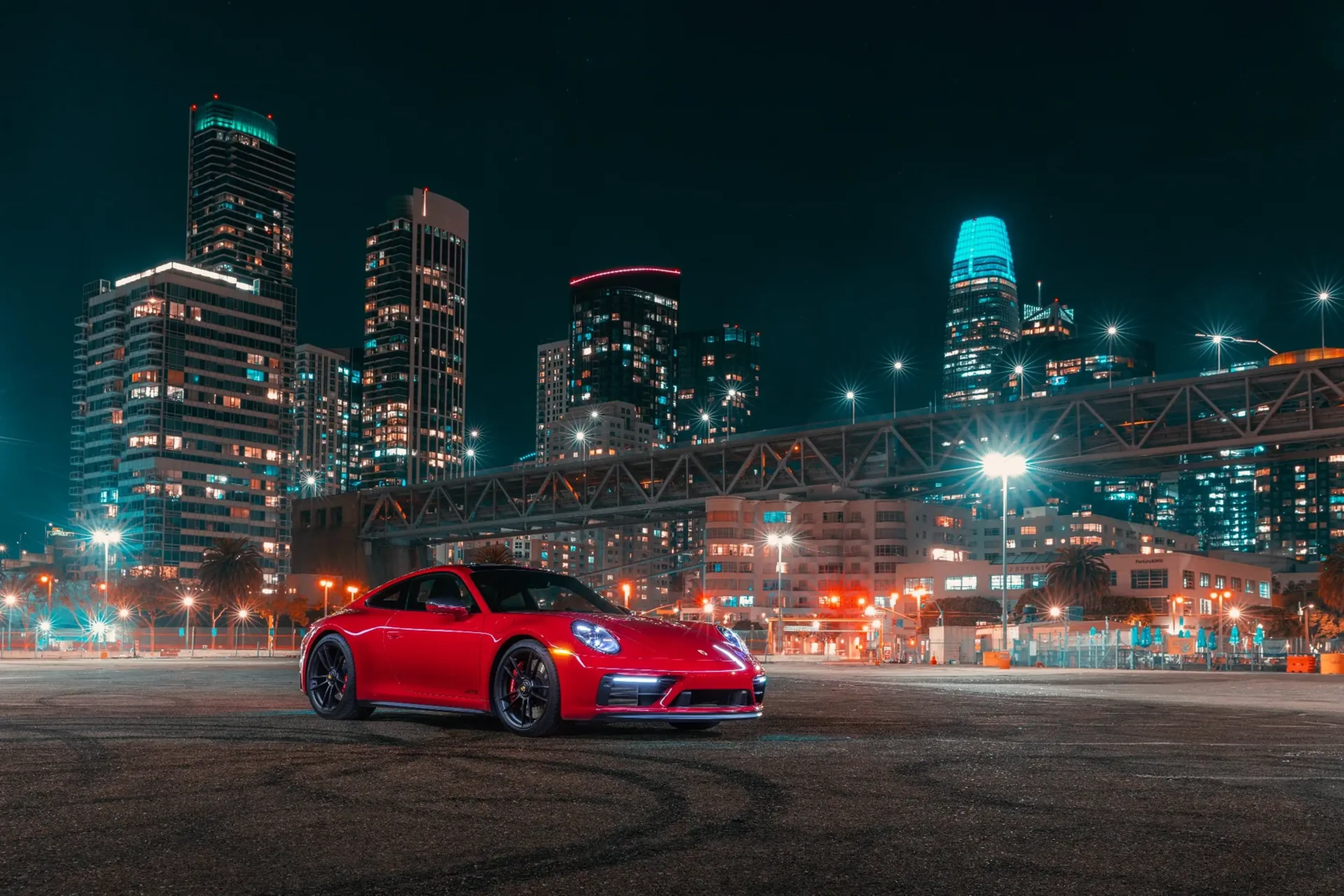 A red sports car parked in an empty lot at night, with illuminated city skyscrapers and a bridge in the background.