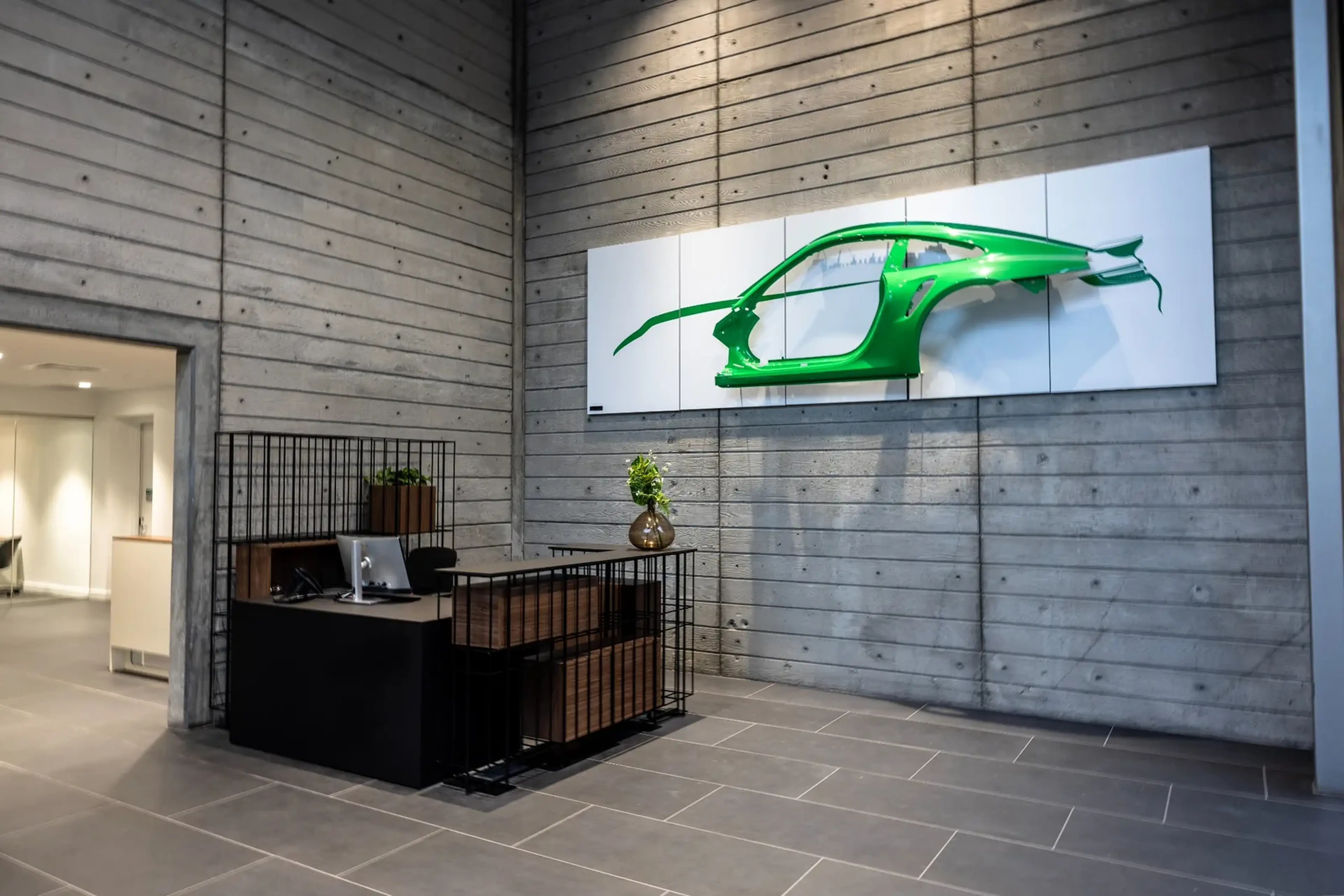 Modern office lobby with concrete walls, a sleek black reception desk, and a green car frame art piece mounted on the wall.