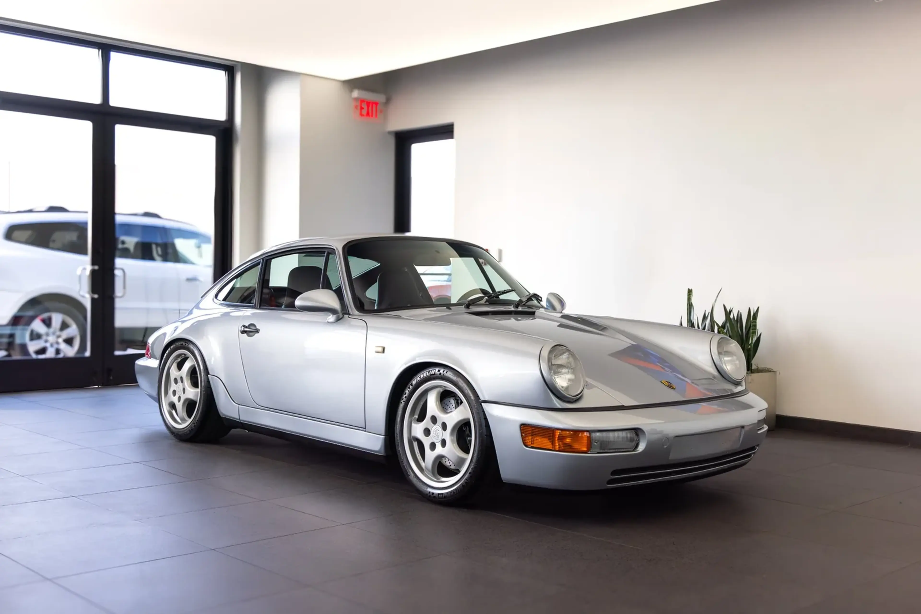 Silver classic sports car with a sleek, vintage design, parked indoors beside a large window and potted plants.