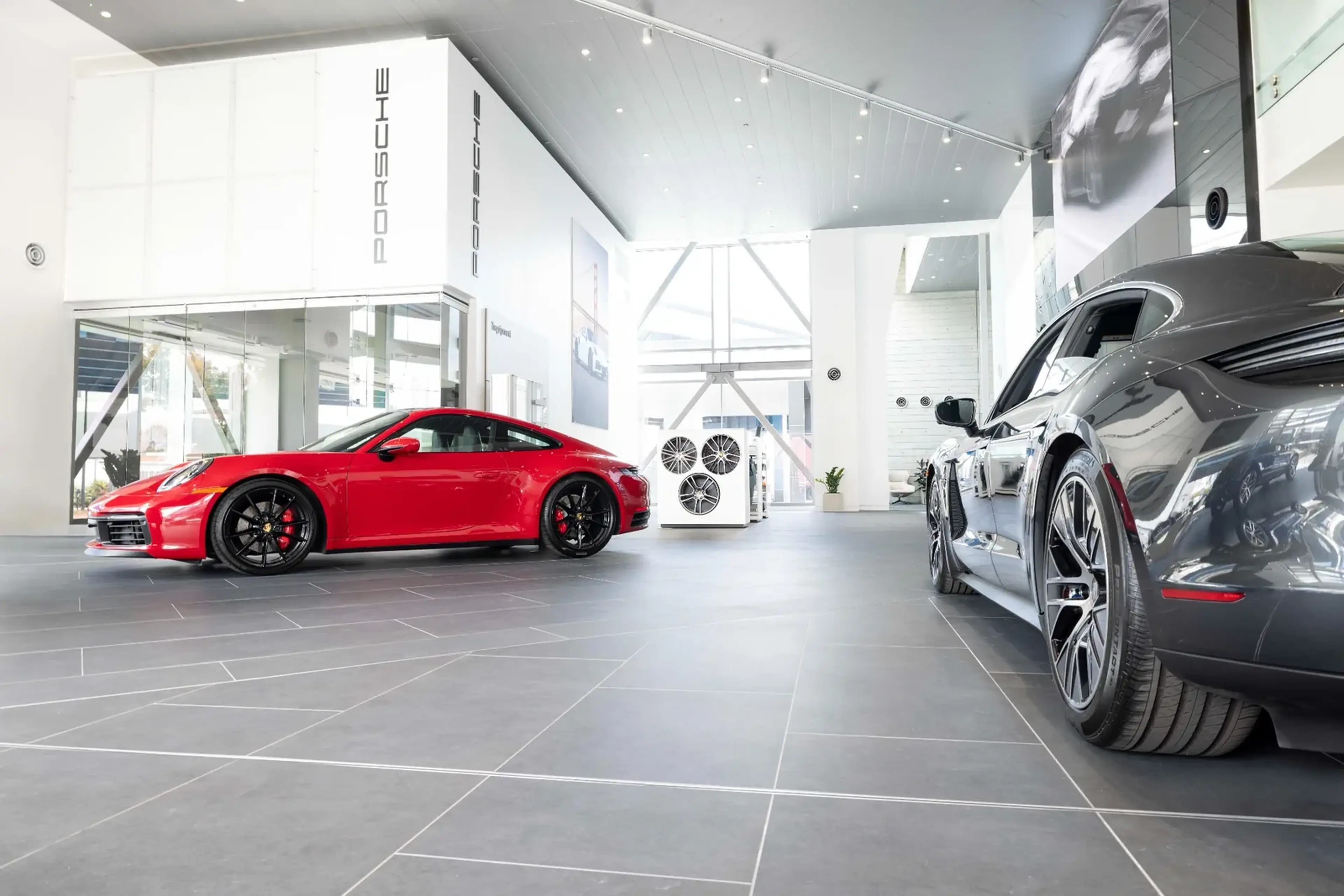 Two sports cars are displayed inside a modern showroom with large windows, featuring a red car on the left and a gray car on the right.