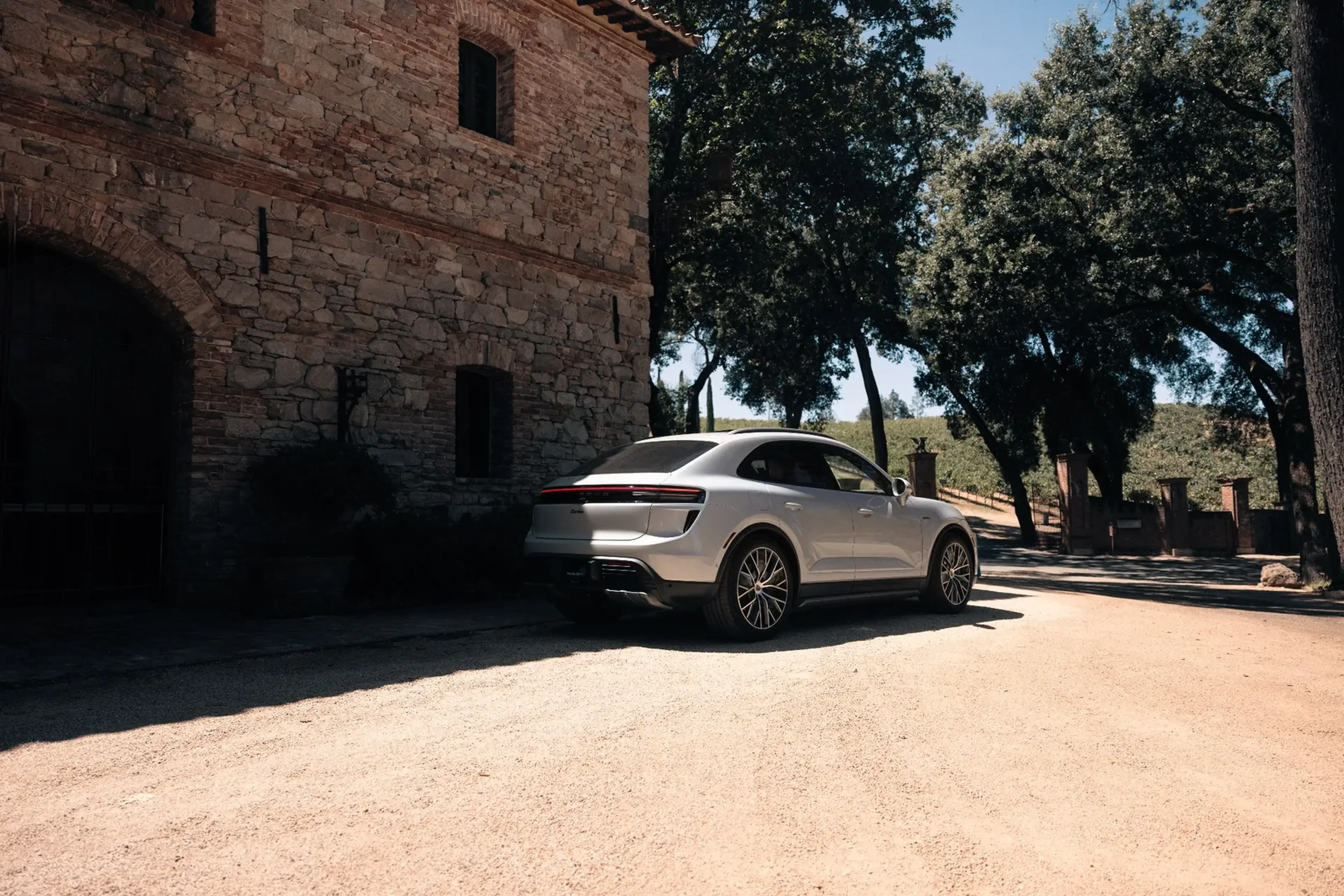A silver luxury car parked on a sunlit dirt path beside a rustic stone building, surrounded by greenery and trees.