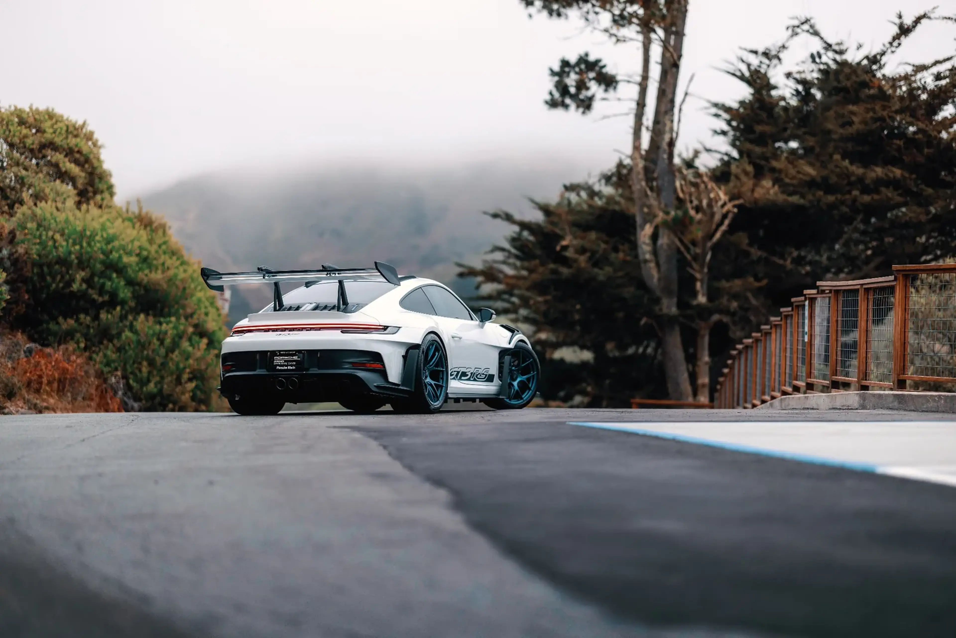 A white sports car on a scenic road with trees and fog in the background, featuring a large rear spoiler and sleek design.