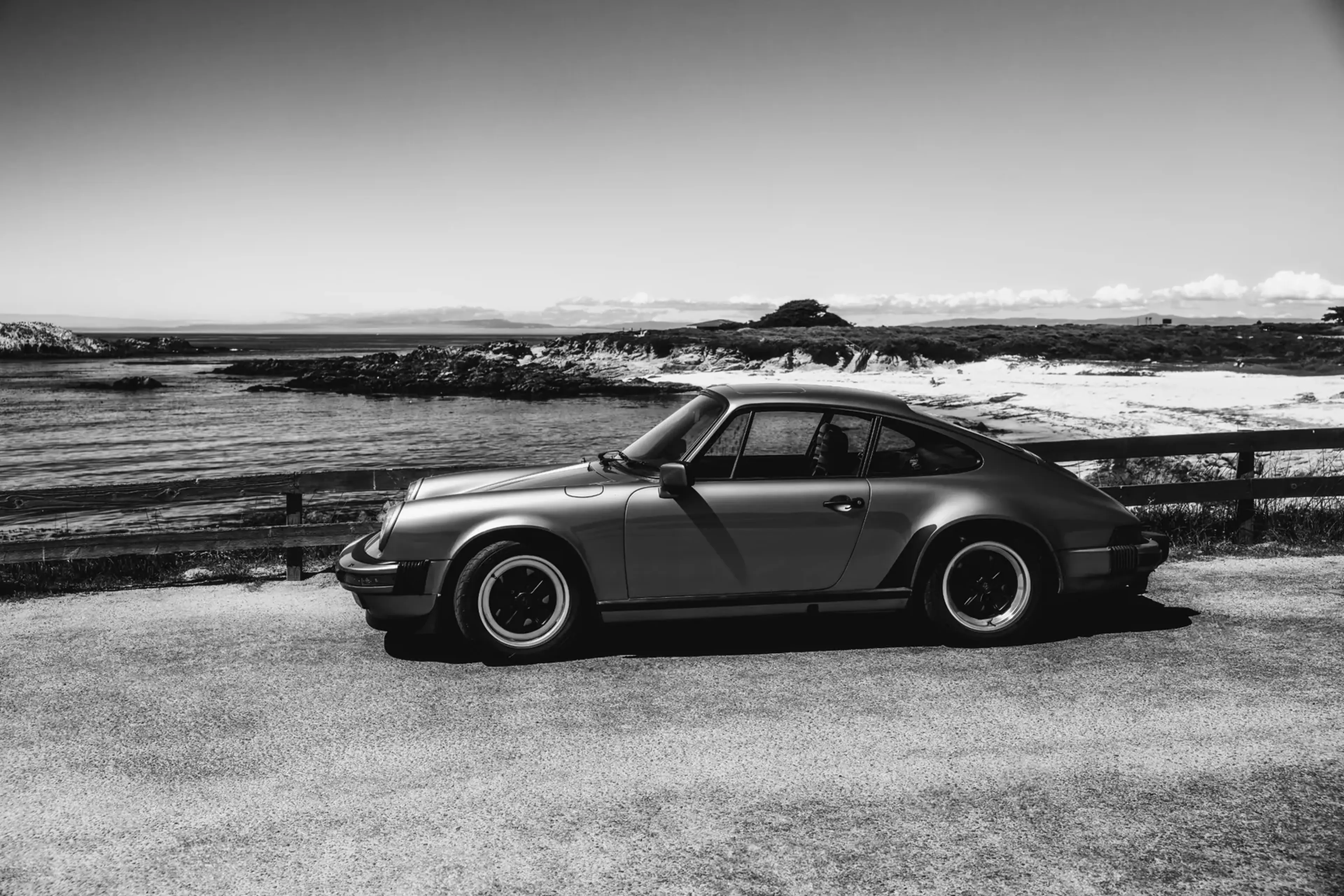 Black and white image of a classic car parked by the ocean with rocky shoreline and clear sky in the background.